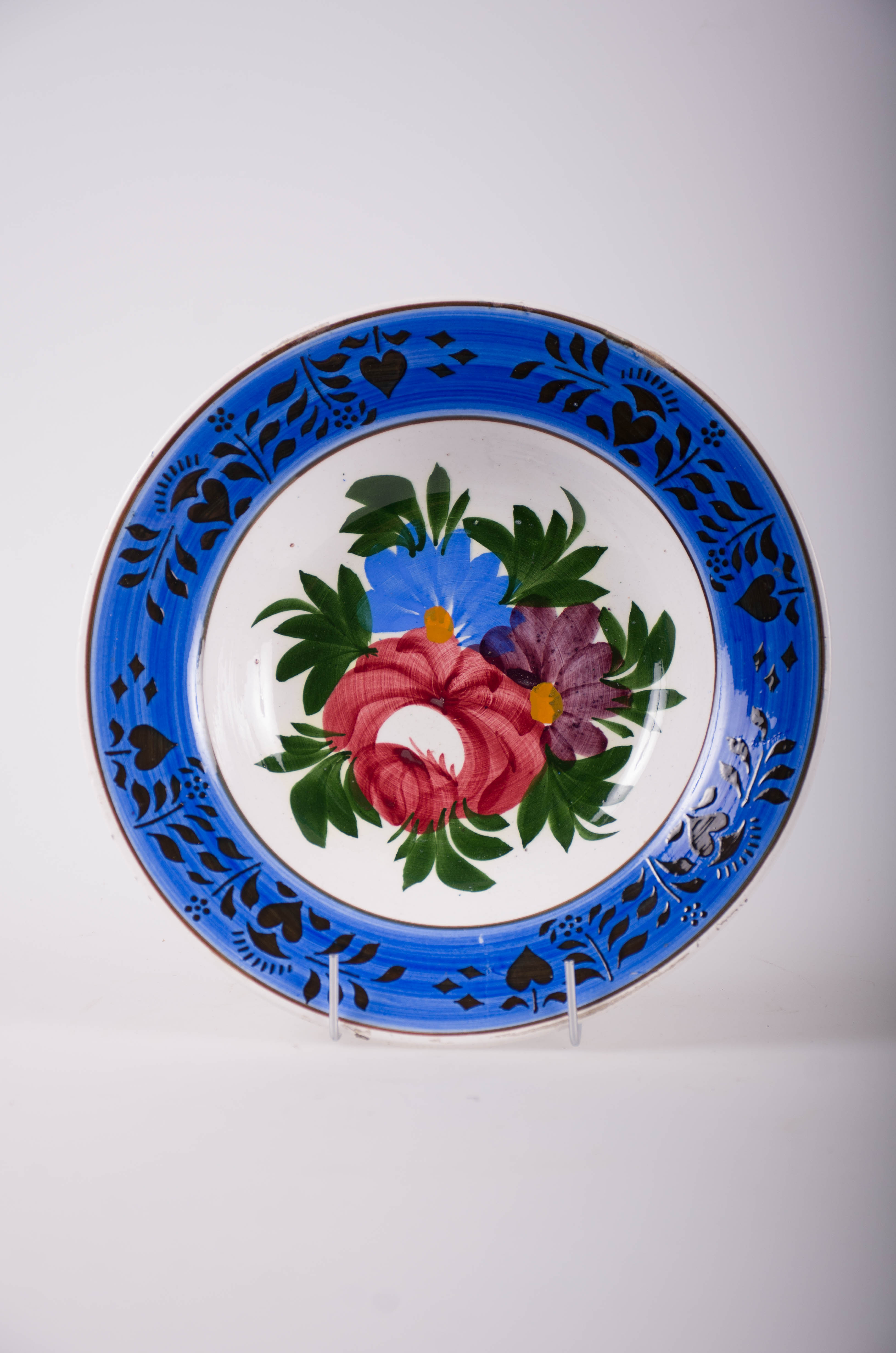 Farfurie de formă tronconică, adâncită; pe fund are găuri pentru agățare. În centru este decorată cu motive fitomorfe și florale; pe margine este decorată cu motive geometrice (linie, romb, punct): două cercuri concentrice încadrează patru registre de flori stilizate. Pe fund are marcă. Cromatică: fond: alb; motive: albastru; verde; negru; vișiniu; galben.