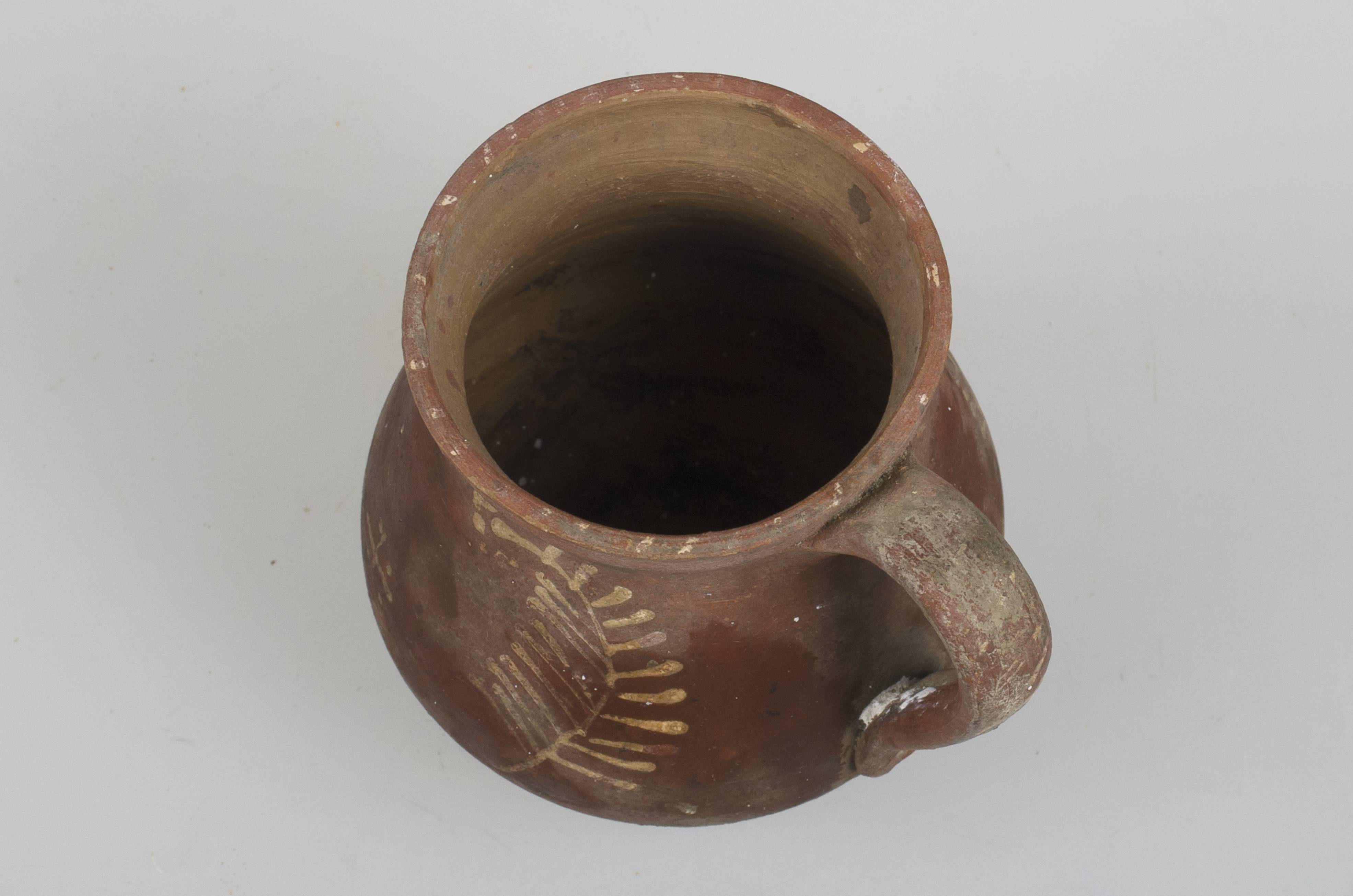 Vas ceramic de formă bitronconică cu baza dreaptă și plată, având gâtul scurt, cu gura largă și buza înălțată. Piesa prezintă o toartă, care pornește de sub buză și care se unește cu vasul în zona diametrului maxim. Este smălțuit în exterior parțial. Vasul este decorat cu motive vegetale: trei crenguțe de brad si o cruce de mici dimensiuni incadrata de patru puncte. Toarta prezintă un ornament format din mai multe linii paralele orizontale oblice. Cromatică: fond: cărămiziu; motive: alb.