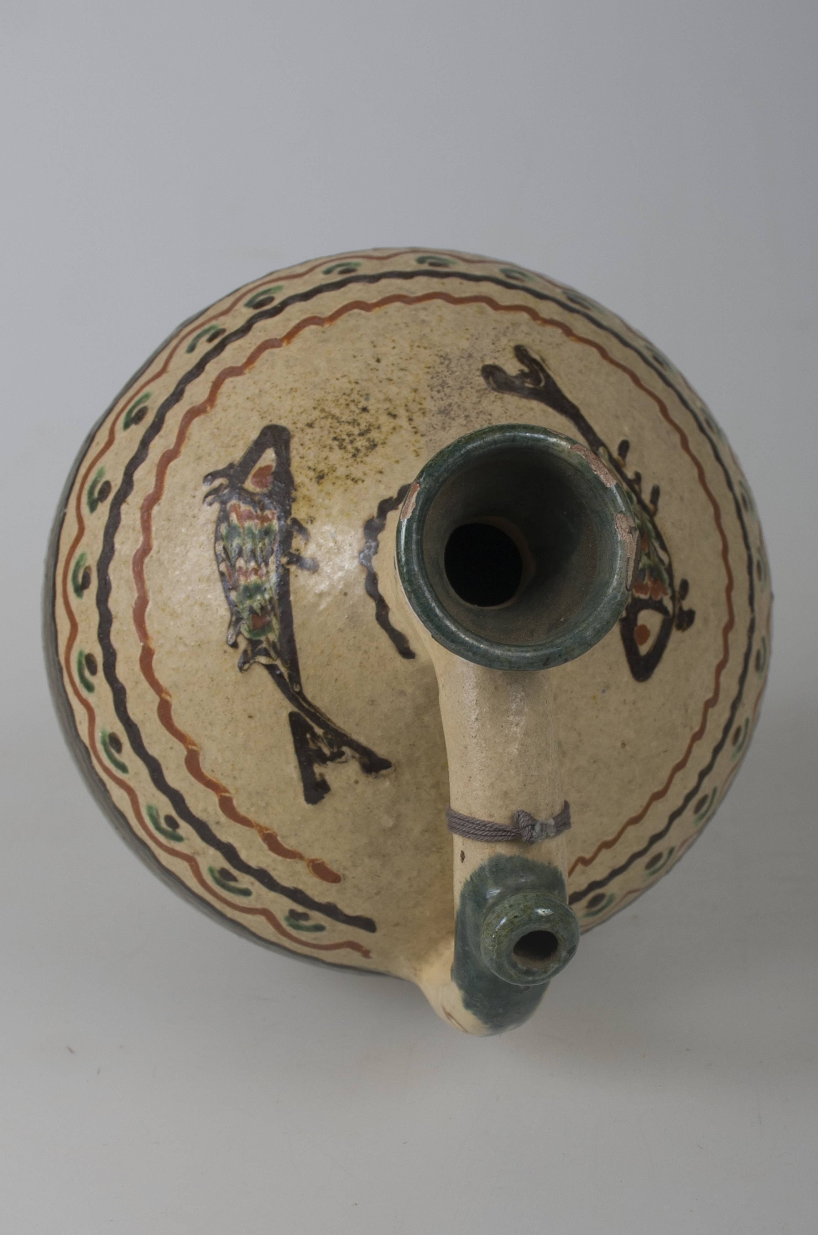Vas ceramic de formă cilindrică, cu baza plată și bombat în zona „pântecelui”, are gâtul și gura reduse ca diametru. O toartă pornește dinspre gât, spre diametrul maxim al vasului, având la jumătatea lungimii o „țâță”, care permitea servirea conținutului lichid. Vasul a fost angobat - angobă verde-închis în jumătatea inferioară și angobă gălbuie, în jumătatea inferioară. Peste stratul de angobă gălbuie a fost realizat decorul compus din linii paralele șerpuite și în zig-zag. Pe „umerii” vasului au fost pictați doi pești, deasupra cărora a fost pictată o linie în valuri, care închide decorul pictat. La gura vasului și „țâță”, recipientul prezintă pete de culoare verzuie. Cromatică: verde; crem; maro-roșcat; maro-închis.