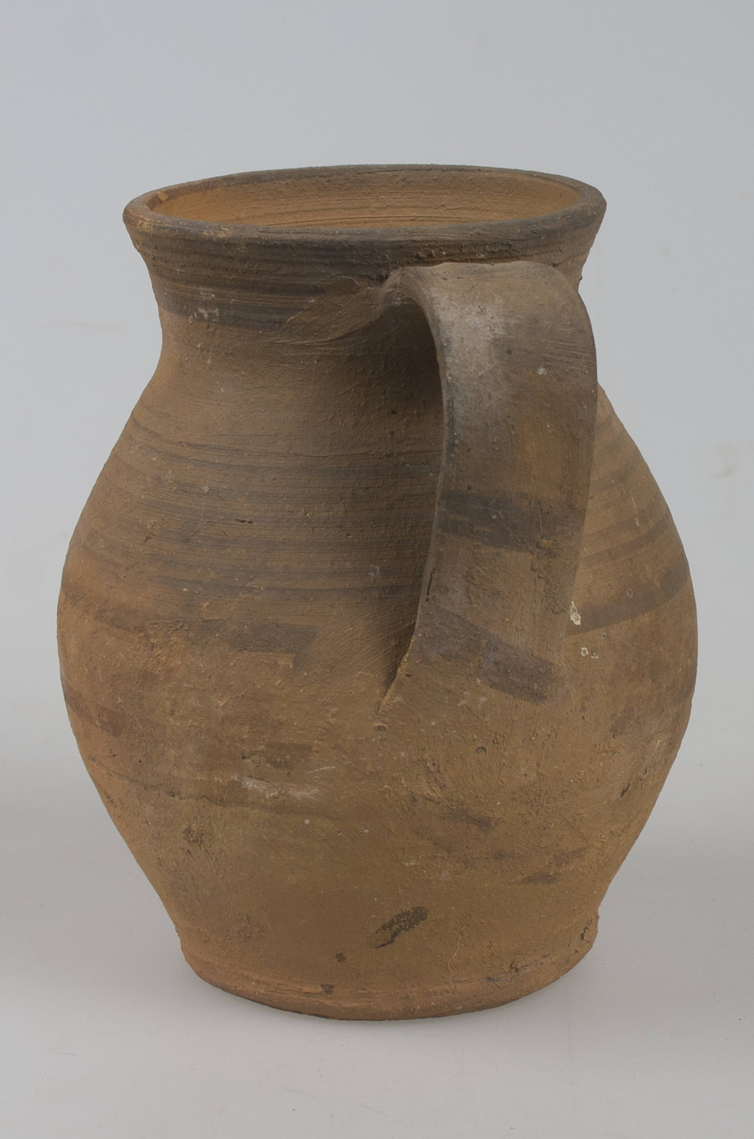 Vas ceramic de formă bitronconică, cu baza dreaptă și plată, având gâtul înalt și profilat, cu gura largă și buza tăiată drept. Piesa prezintă o toartă, care pornește de sub buză și care se unește cu vasul în zona diametrului maxim al acestuia. Corpul nesmălțuit al vasului este decorat în jumătatea superioară cu motive geometrice: linii concentrice decorează umerii și gâtul vasului, toarta fiind ornamentată la rândul ei, cu mai multe linii ușor oblice paralele. Cromatică: fond: maro deschis; motive: negru.