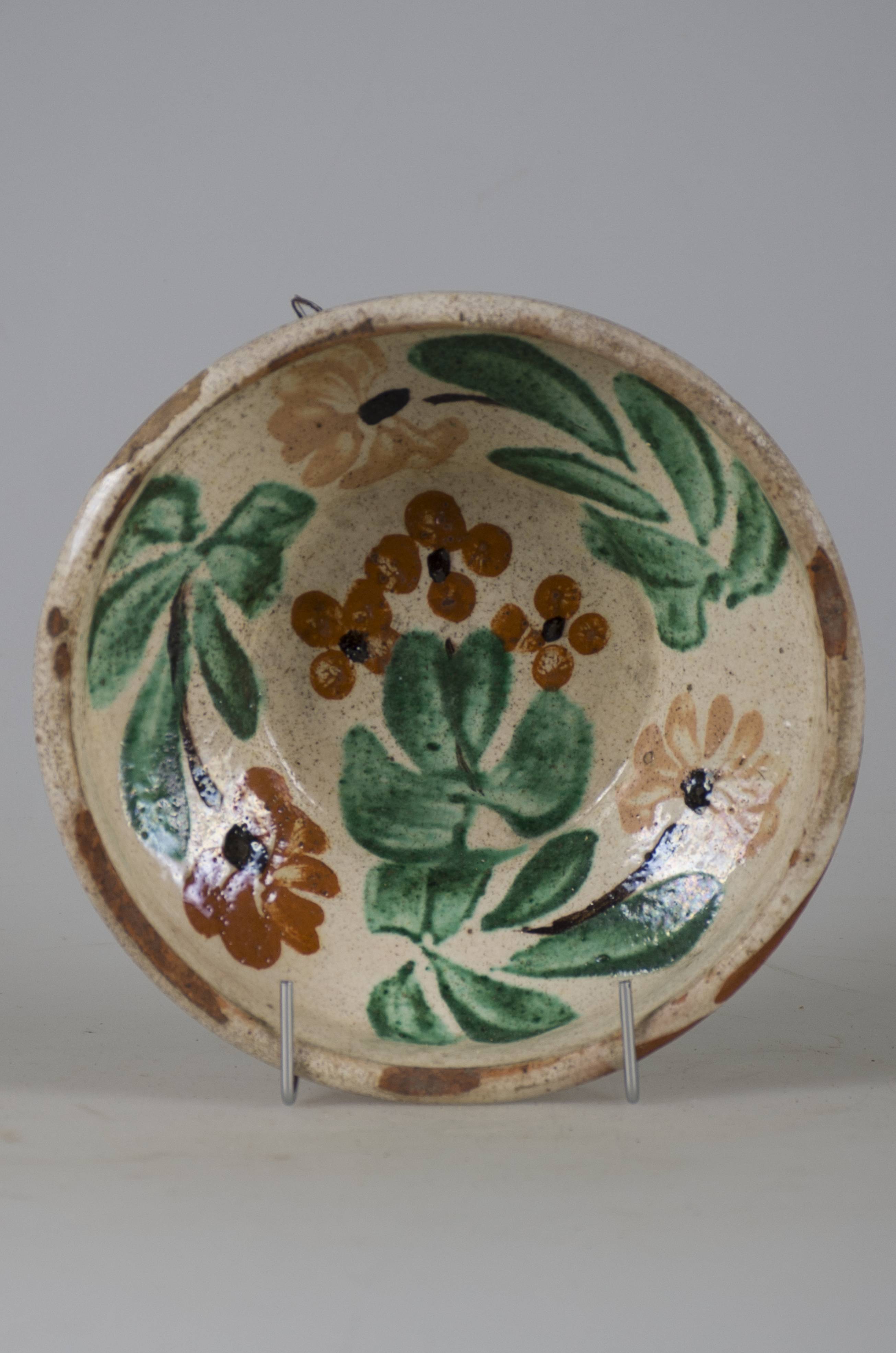 Vas ceramic smălțuit în interior, de formă tronconică, cu marginile puțin ridicate și fundul plat. Pe fond alb prezintă decorații realizate prin pictare cu pensula, ilustrând trei flori, două albe și una roșie, dispuse circular și despărțite de frunze verzi, plasate perimetral pe pereții interiori al vasului. Pe fundul farfuriei avem un grup de trei flori roșii, de mai mici dimensiuni cu frunze verzi. Florile au tulpini negre și câte un punct negru în centru. Pe partea din spate, la bază, prezintă două orificii pentru suspendare. Cromatică: fond: alb; motive: alb; roșu; verde; negru