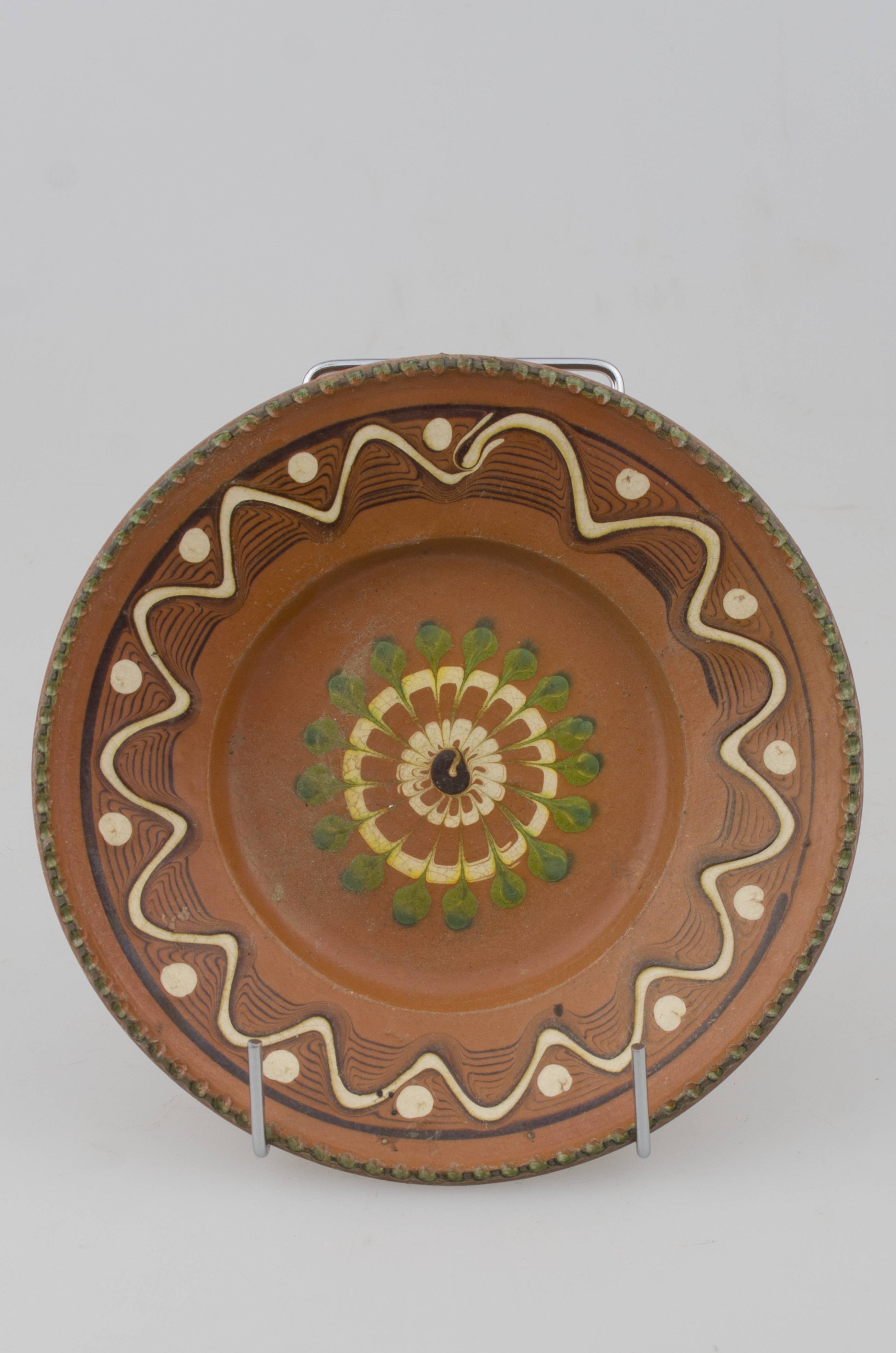 Farfurie cu pereții rotunjiți, marginea oblică lată și buza crestată. Prezintă decor geometric pe margine: linii frânte și curbe; pete de culoare; central avem o floare stilizată. Cromatică: fond: maro; motive: verde; alb.