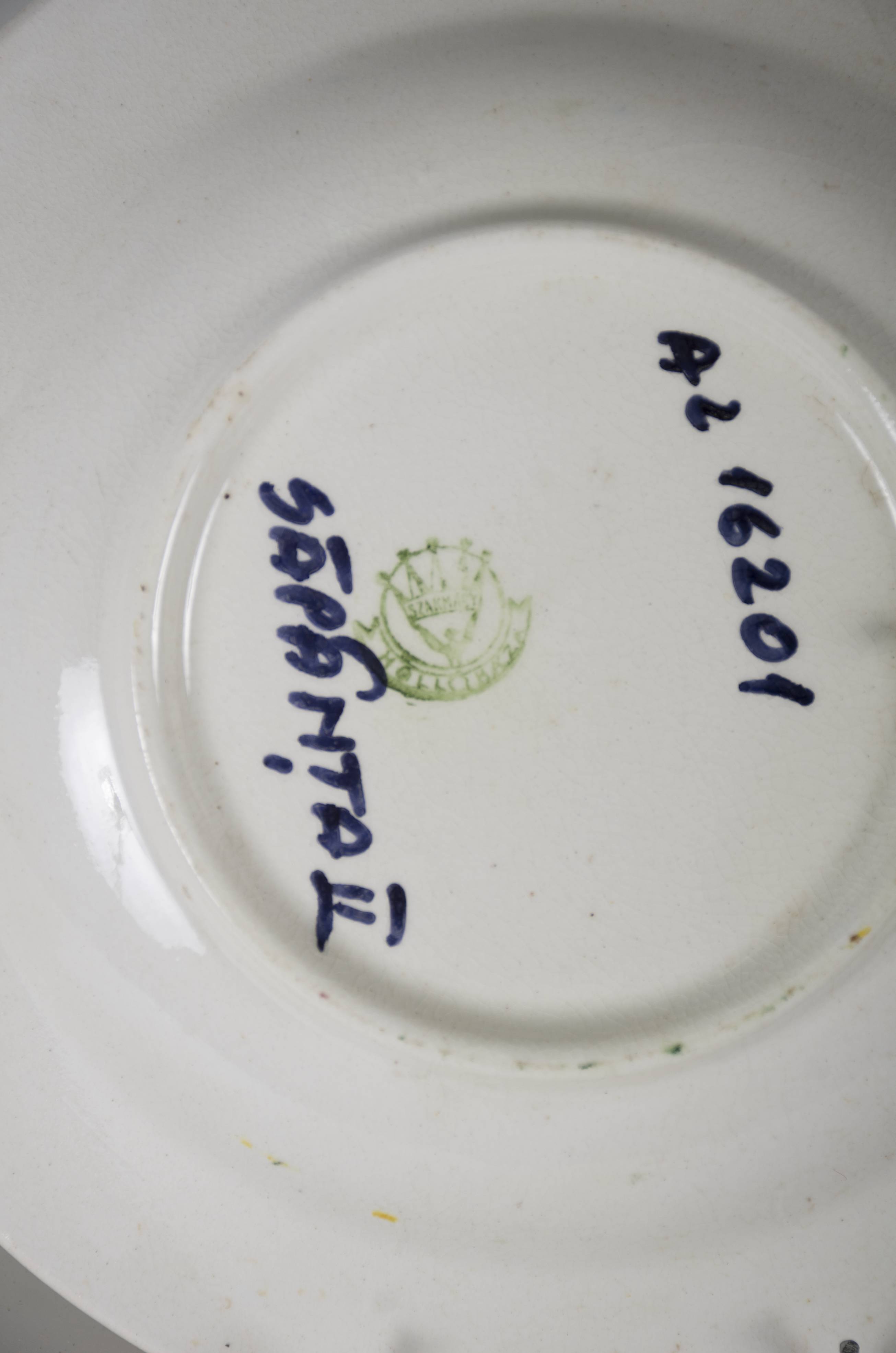 Vas ceramic smălțuit, de formă tronconică, cu marginile puțin ridicate și fundul plat. Pe fond alb prezintă decorații în registru floral-vegetal ilustrând patru flori poziționate central: două cu petale roșii, celelalte de culoare galbenă și albastră. Compoziția se completată cu frunze verzi, tulpini maro și trei flori de mici dimensiuni cu petale roșii. Pe marginea farfuriei avem o decorație circulară dantelată. Pe spate, la bază, prezintă două orificii pentru suspendare. Cromatică: fond: alb; motive: roșu; galben; albastru; verde.