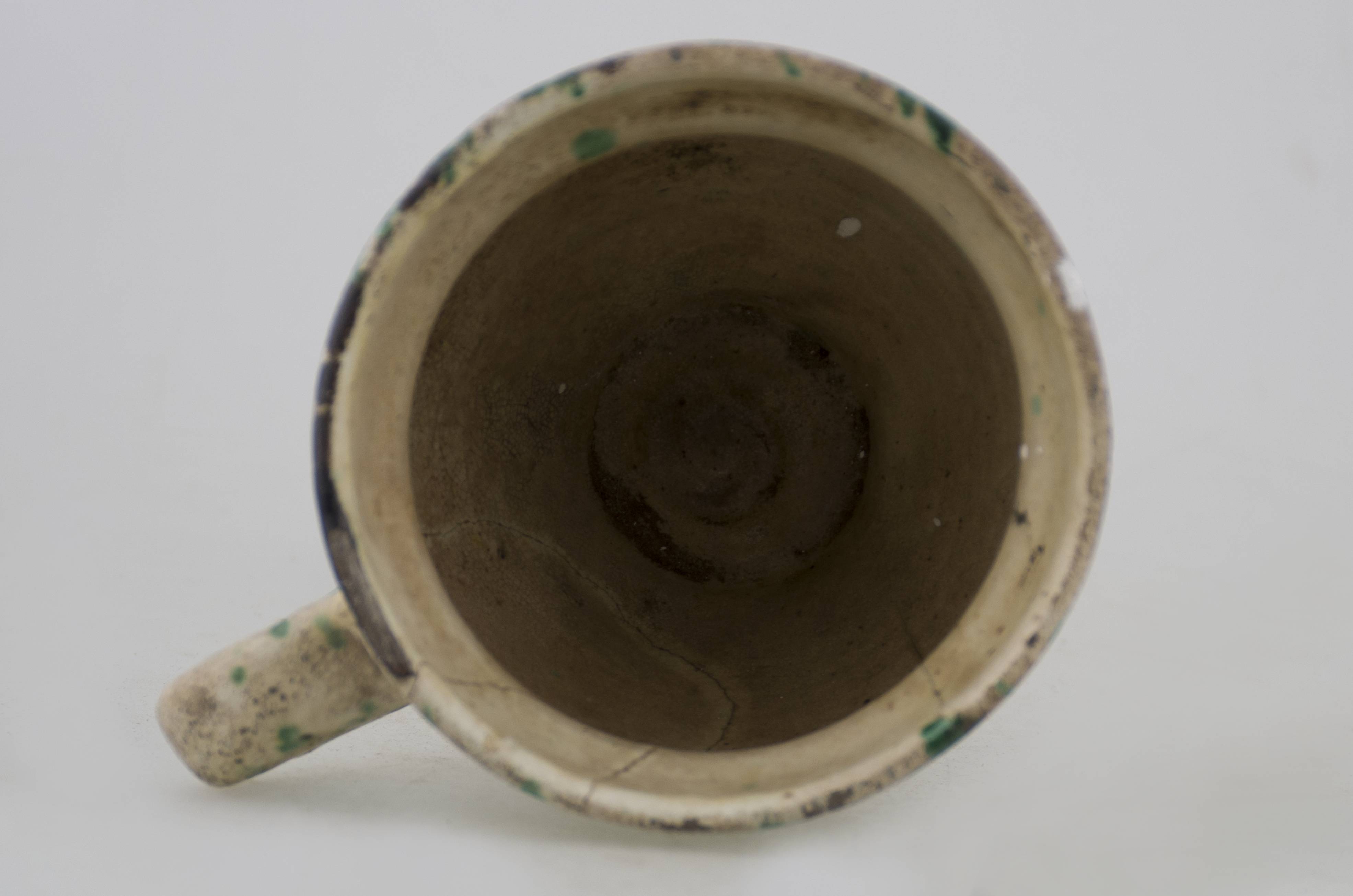Recipient ceramic bitronconic, de mici dimensiuni, cu gât foarte profilat, gura largă și evazată, mult mai mare decât baza. Prezintă o toartă care pornește de sub buza vasului și se unește cu corpul aproape de bază. Este smălțuită în interior și în exterior (mai puțin baza pe alocuri). Piesa este decorată pe un fundal alb cu pete de culoare verzi realizate cu pensula. Cromatică: fond: alb/crem; motive: verde.