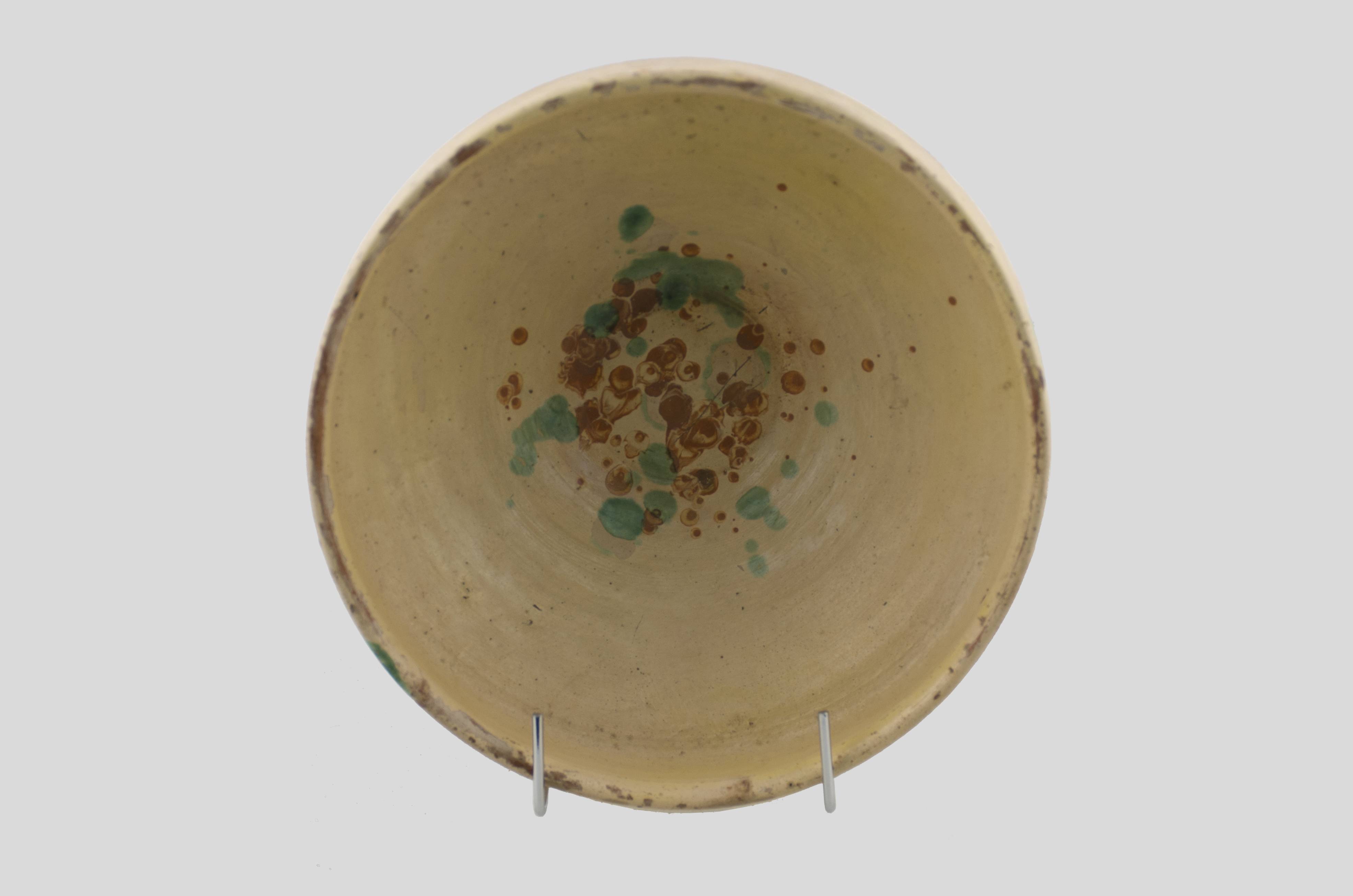 Vas ceramic tronconic, de dimensiuni reduse ce prezintă baza plată, pereții oblici și buza înălțată și ieșită înafară. Vasul este decorat cu motive vegetale stilizate realizate din stropi amplasate în centru. Baza exterioară a recipientului ceramic are incizate inițialele meșterului „ I. B.”. Cromatică: fond: alb-galbui; motive: roșu; verde.