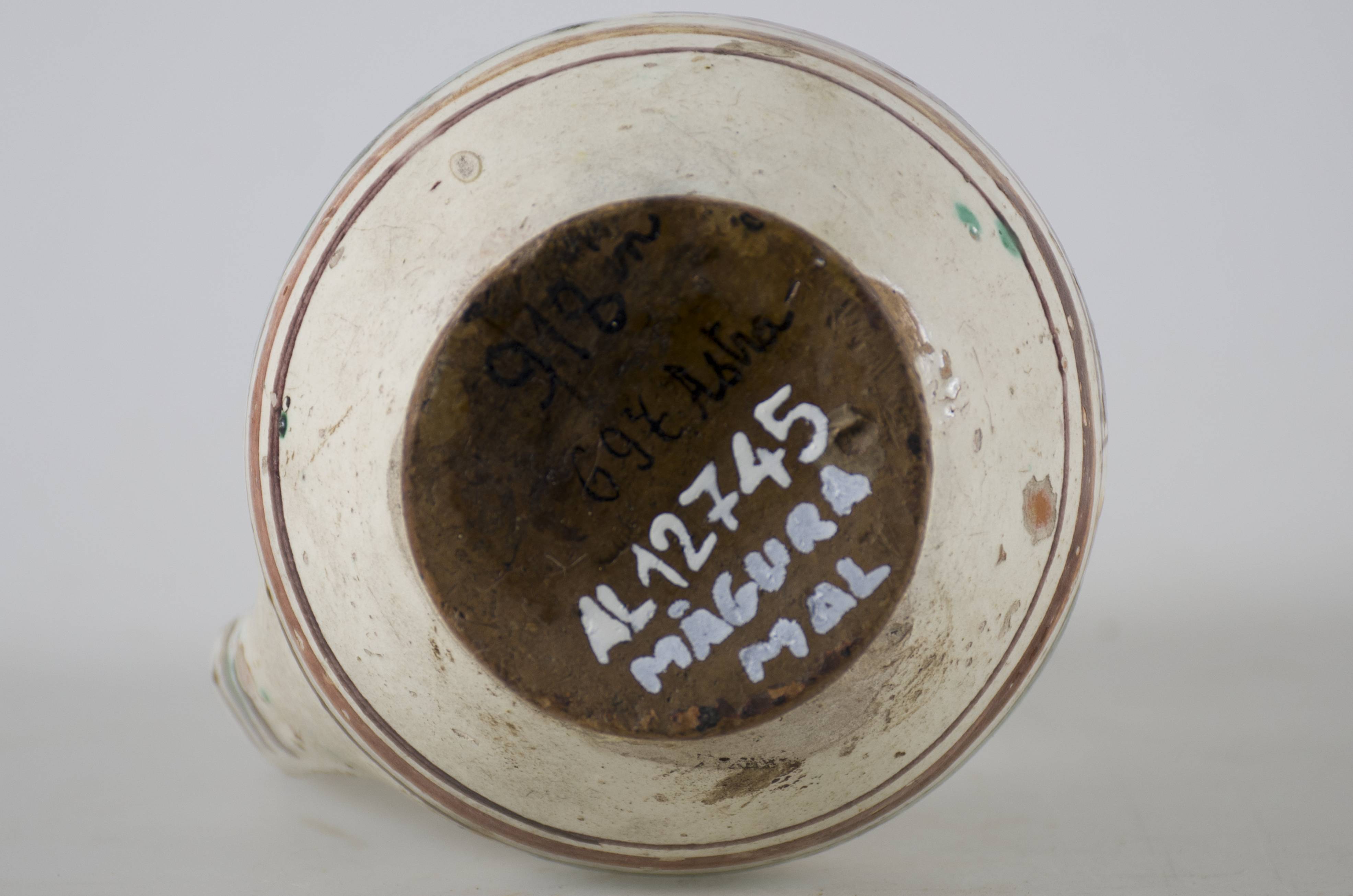 Vas ceramic smălțuit de mici dimensiuni, piriform, cu baza plată și bombat în zona „pântecelui”. Prezintă o toartă ce pornește din zona gâtului redus ca dimensiuni și se termină în zona diametrul maxim al vasului. Decorul vasului este compus din trei registre picturale. În primul registru au fost pictate două flori albe pe fundal verde și o a treia pe fundal maro, fiind despărțite prin linii de culoare galbenă. Cel de-al doilea registru a fost decorat cu motive geometrice stilizate, linii și triunghiuri afrontate de culoare galbenă, pe fundal de culoare verde. Ultimul registru a fost decorat cu linii de culoare maro și galben, asemenea cu trei brazi și înconjurate de câte două frunze de culoare verde. Toate cele trei registre picturale sunt încadrate de câte trei linii concentrice (două subțiri și una lată) de culoare maro-închis și maro-roșcat. Pe toartă este decorat cu linii paralele orizontale. Cromatică: fond alb; motive: galben; verde; maro.
