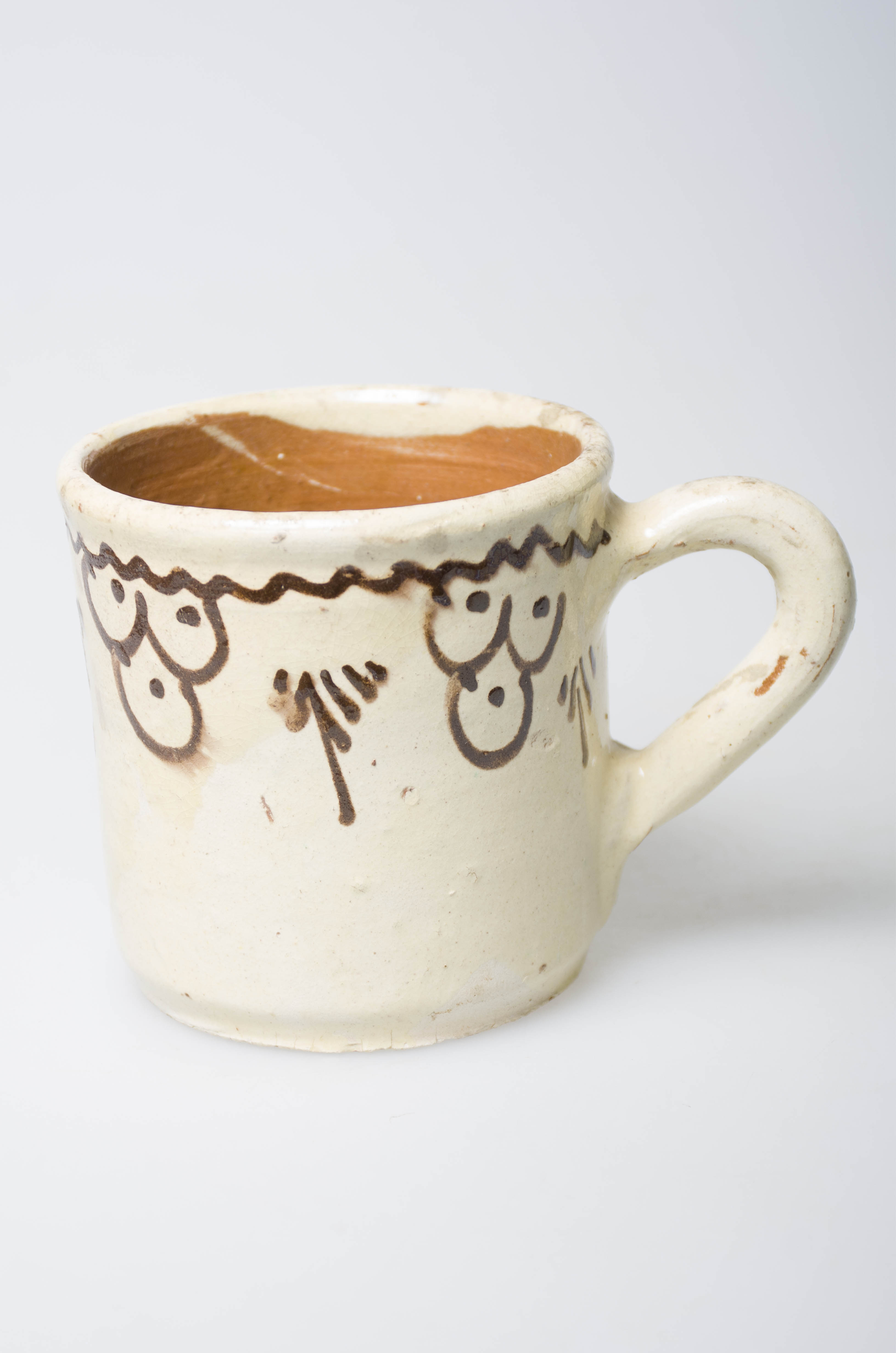 Ceașca este confecționată din ceramică, având forma cilindrică. Aceasta este prevăzută cu o toartă mare. Decorul este pictat: sub buză există o linie ondulată, cu grupe de 3 semicercuri, cu câte un punct în fiecare dintre acestea. Obiectul este smălțuit. Cromatica: fondul este alb iar motivele sunt pictate cu maro.