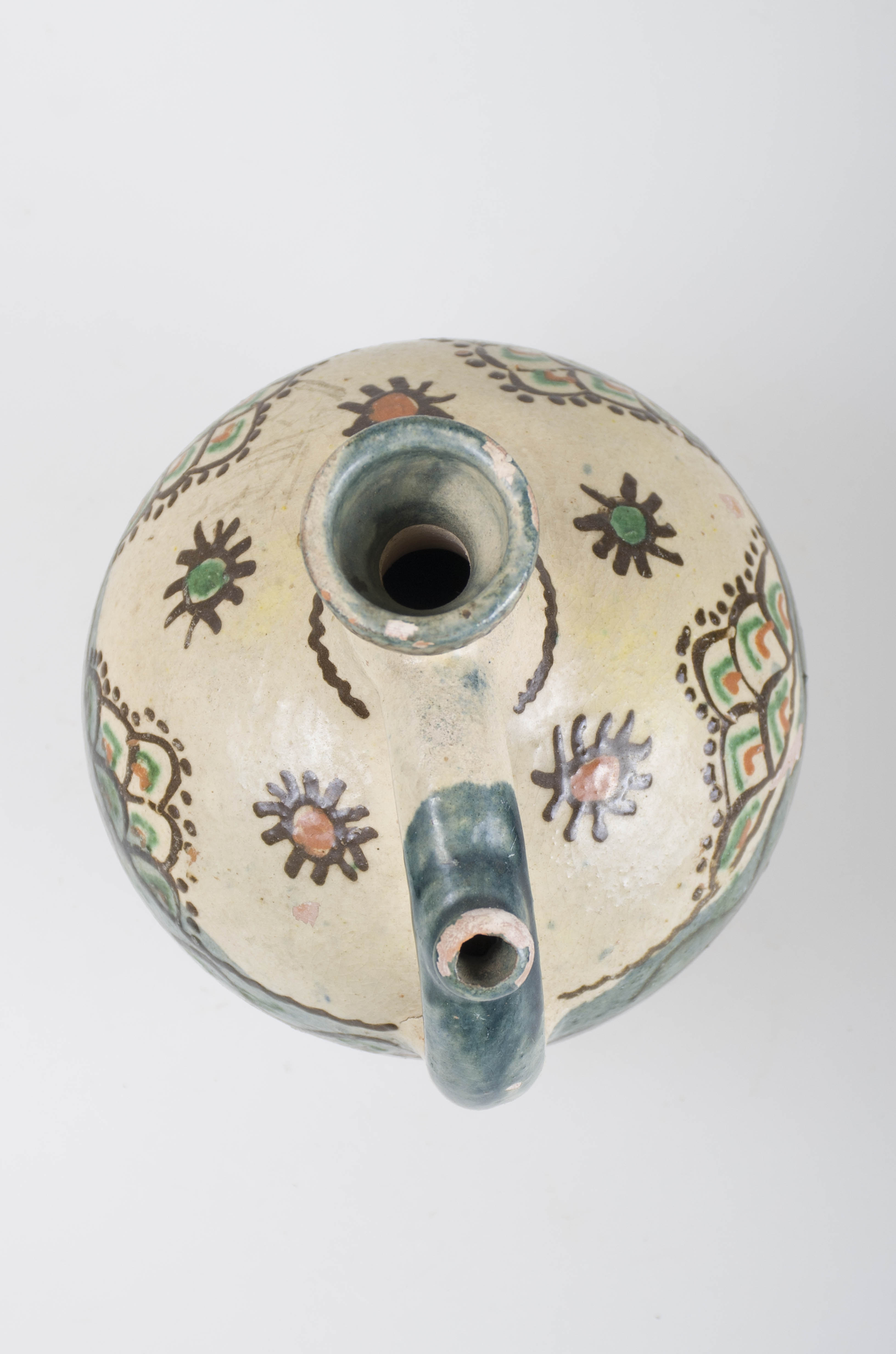 Vas ceramic piriform, smălțuit; cu gât înalt cu guleraș din care coboară spre partea mediană o toartă, prevăzută cu "țâță", pentru scurgerea lichidului. Acesta este decorat în mai multe registre cu diferite motive geometrice și vegetale: în zona mediană întâlnim o line vălurită încadrată între două linii meandrice, urmată de semicercuri tricolore puse unele peste altele, alcătuind un triunghi înconjurat de puncte mărunte; pe umeri sunt pictate trei flori cu petale subțiri și o linie frântă, meandrică; la gura recipientului este pictată o linie vălurită. Obiectul este smălțuit pe toartă, gură și în partea inferioară a vasului. În partea superioară, prezintă angobă albă. Cromatică: fond: alb; verde; motive: cărămiziu, verde; maro închis.