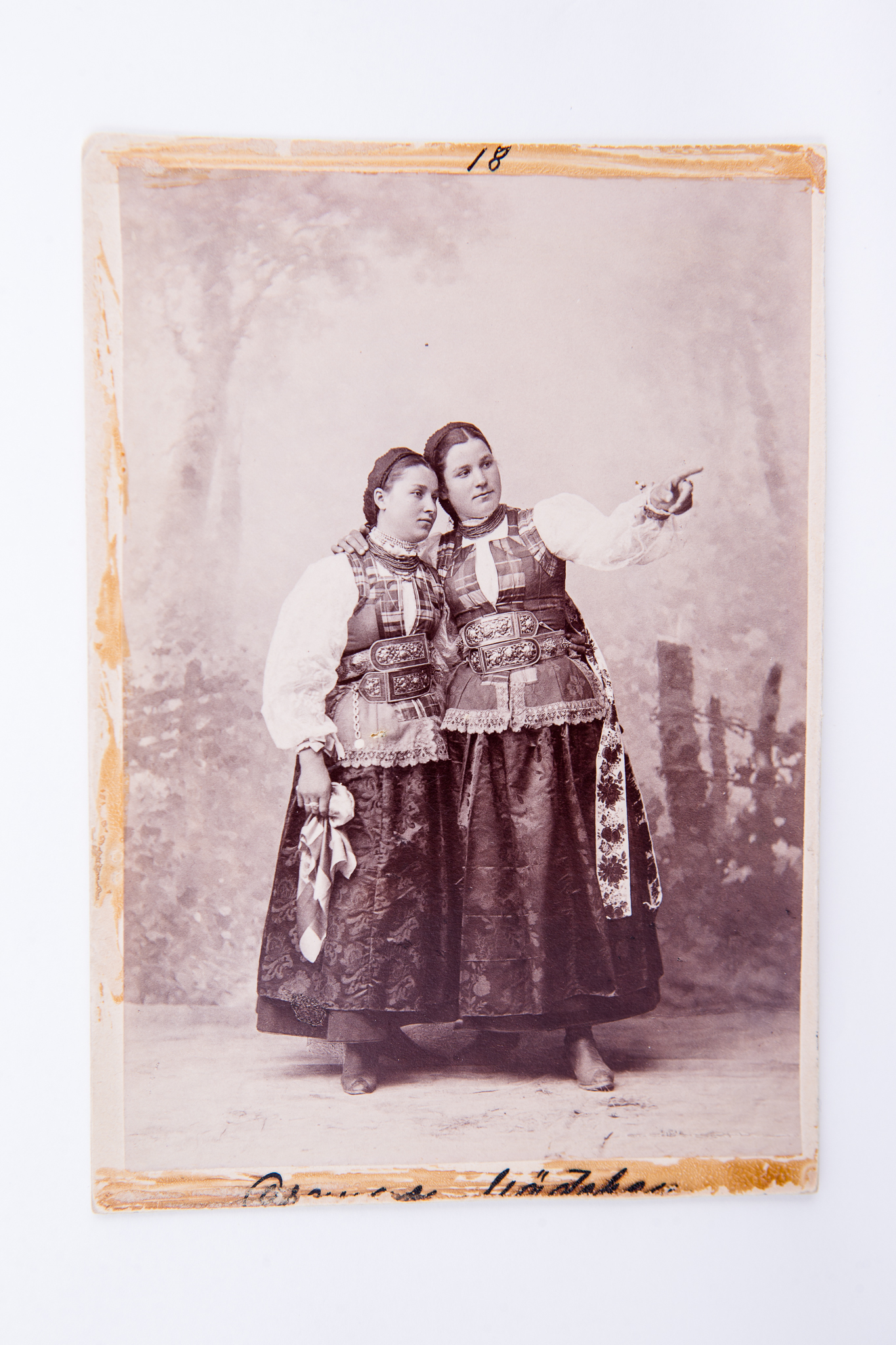 Tinere în costume populare de sărbătoare de ceangăi. Fotografie de atelier cu fundal pictat. Pe carton, în partea de jos, însemnare manuscrisă incomplet vizibilă. Pe trei laturi fotografia prezintă urme de lipire / dezlipire de pe un alt suport. Deasupra hârtiei sensibilizate, pe carton, însemnare manuscrisă cu stiloul: "18". Pe verso marca atelierului fotografic cu doi grifoni și monograma autorului și text de reclamă în limba germană: "Fotograf Leopold Adler / Kronstadt / Purzengasse / Vervielfältigung vorbehalten / Nachbestellung werden selbst nach Jahren ebenso sorgfälting ausgeführt / Kuhle und Miksche / Wien" (Reproducerea rezervată / Comenzile ulterioare vor fi executate chiar și după ani cu aceeași acuratețe.) Însemnări manuscrise cu creionul ("Fete de Ceangău"), număr inventar MCPR 638.