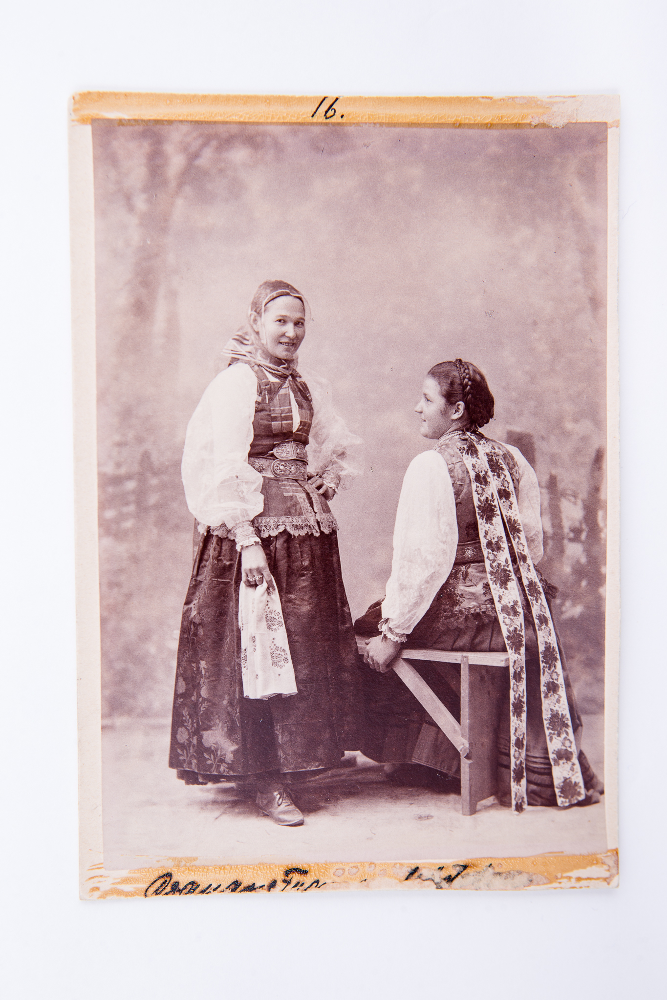 Două femei în costume populare de ceangăi. Una dintre femei este așezată pe o bancă, iar cealaltă femeie stă în picioare lângă bancă. Fotografie de atelier cu fundal pictat și elemente de recuzită (banca). Pe carton, în partea de jos, însemnare manuscrisă incomplet vizibilă. Pe trei laturi fotografia prezintă urme de lipire / dezlipire de pe un alt suport. Deasupra hârtiei sensibilizate, pe carton, însemnare manuscrisă cu stiloul: "16.". Pe verso marca atelierului fotografic cu doi grifoni și monograma autorului și text de reclamă în limba germană: "Fotograf Leopold Adler / Kronstadt / Purzengasse / Vervielfältigung vorbehalten / Nachbestellung werden selbst nach Jahren ebenso sorgfälting ausgeführt / Kuhle und Miksche / Wien" (Reproducerea rezervată / Comenzile ulterioare vor fi executate chiar și după ani cu aceeași acuratețe). Însemnări manuscrise cu creionul ("Ceangăi - Țara Bîrsei"), număr inventar MCPR 644.