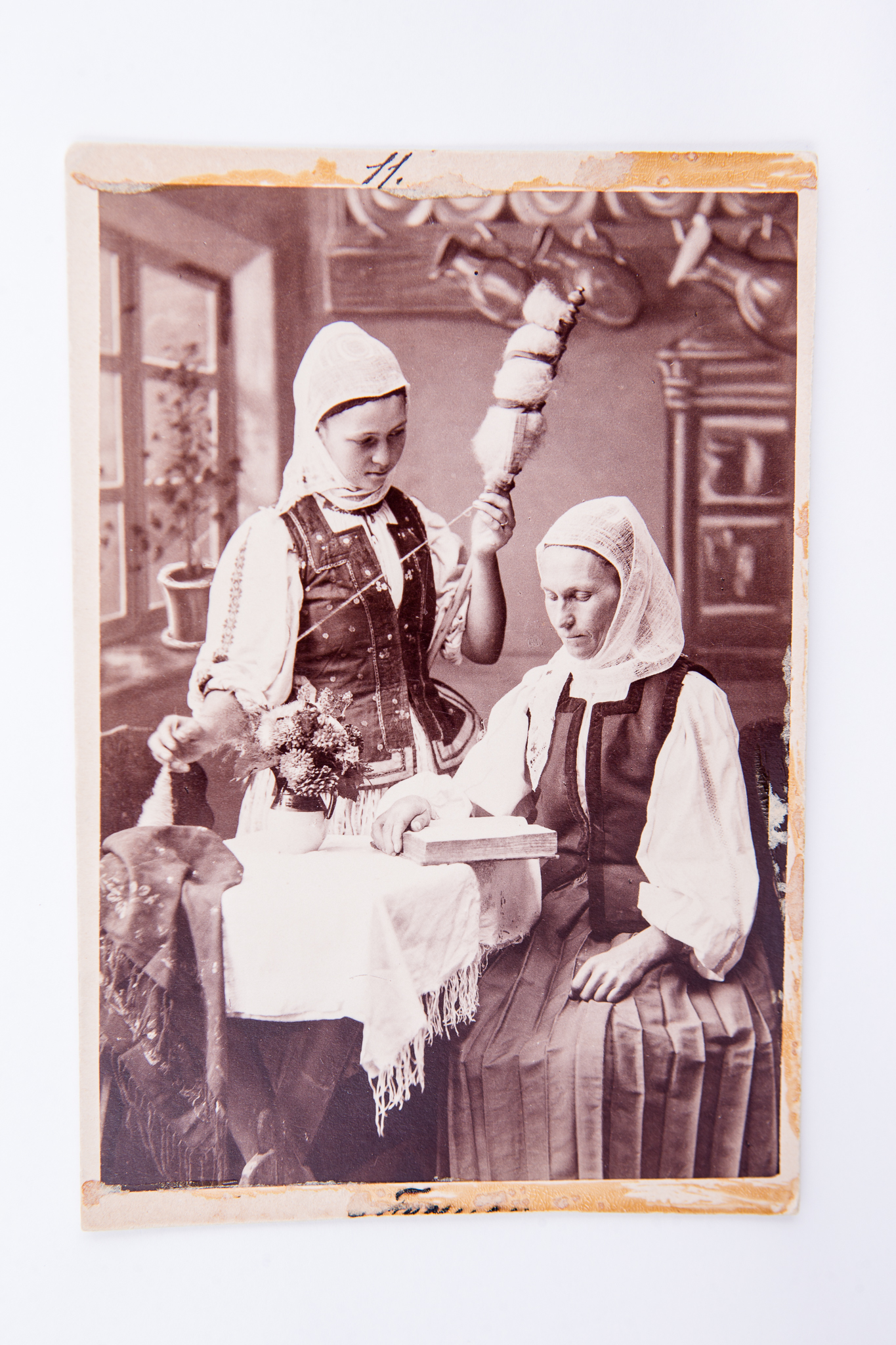 Două femei în costume populare de ceangăi așezate la o masă. Una dintre femei citește, iar cealaltă femeie toarce. Fotografie de atelier cu fundal pictat și elemente de recuzită (scaune, masă, furcă, fuior, fus,flori). Pe carton, în partea de jos, însemnare manuscrisă incomplet vizibilă. Pe trei laturi fotografia prezintă urme de lipire / dezlipire de pe un alt suport. Deasupra hârtiei sensibilizate, pe carton, însemnare manuscrisă cu stiloul: "11.". Pe verso marca atelierului fotografic cu doi grifoni și monograma autorului și text de reclamă în limba germană: "Fotograf Leopold Adler / Kronstadt / Purzengasse / Vervielfältigung vorbehalten / Nachbestellung werden selbst nach Jahren ebenso sorgfälting ausgeführt / Kuhle und Miksche / Wien" (Reproducerea rezervată / Comenzile ulterioare vor fi executate chiar și după ani cu aceeași acuratețe.) Însemnări manuscrise cu creionul ("Ceangăi"), număr inventar MCPR 640.