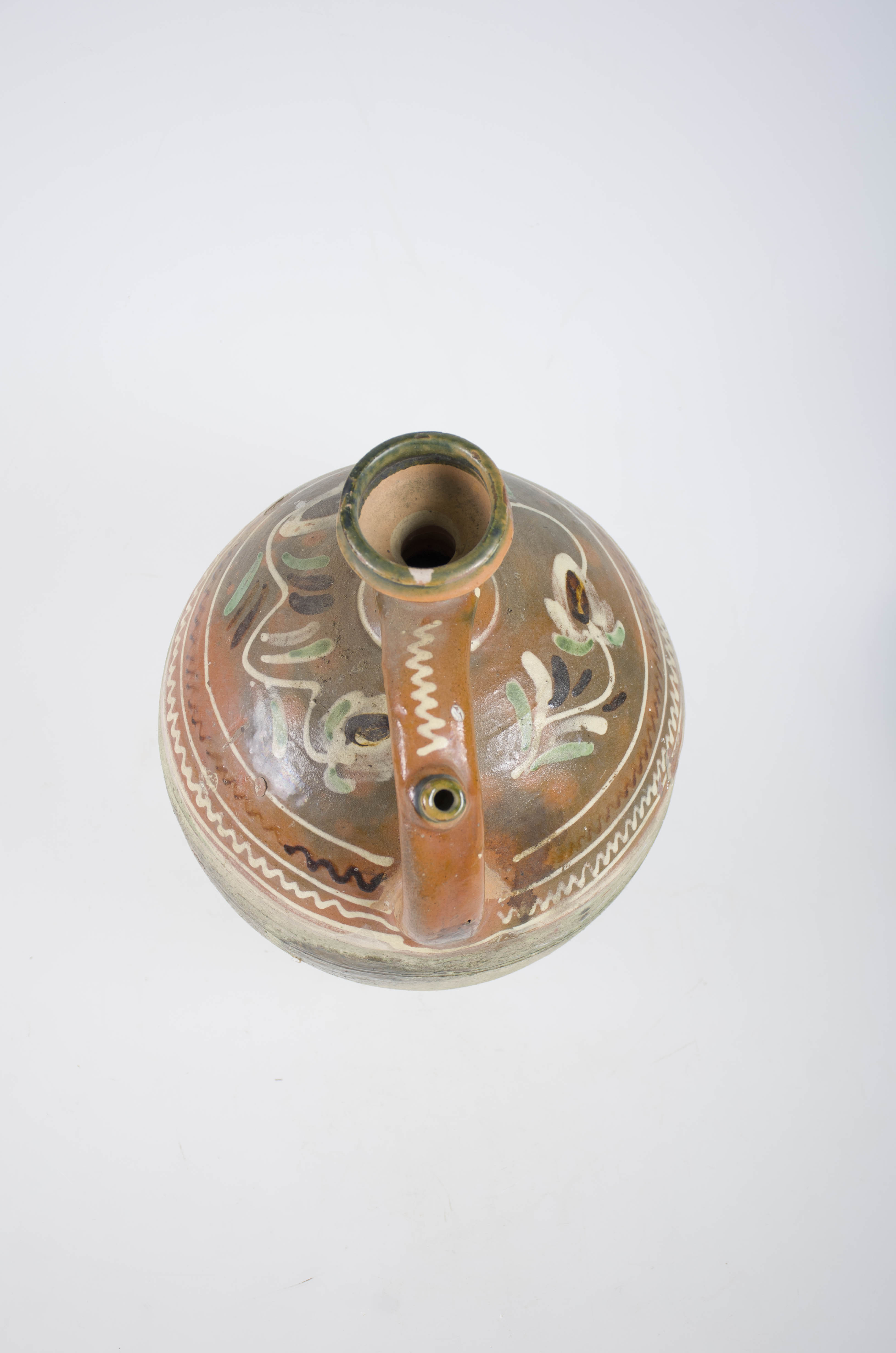 Recipient ceramic, smălțuit, cu pântecul bombat în zona mediană, cu gâtul cilindric și buza dreaptă. Prezintă o toartă tubulară prevăzută cu ”țâță”, pentru scurgerea lichidului. Decorat cu motive geometrice și vegetale, în trei registre pictate cu pensula și delimitate de trei linii circulare subțiri și albe: în primele două registre linii meandrice, iar în cel de al treilea, flori stilizate cu frunze ovoidale, boboci de floare rotunzi și crenguțe. Pe gât, este pictat un șir de puncte albe iar mânerul, până în zona țâței, este decorat cu o linie meandrică albă. Partea inferioară a vasului până la jumătatea pântecului este smălțuită întrun strat foarte subțire de verde deschis, granulat, restul ornamentului fiind pe un fond de smalț cărămiziu. Cromatică: fond: maro, verde deschis, motive: alb, verde, maro.