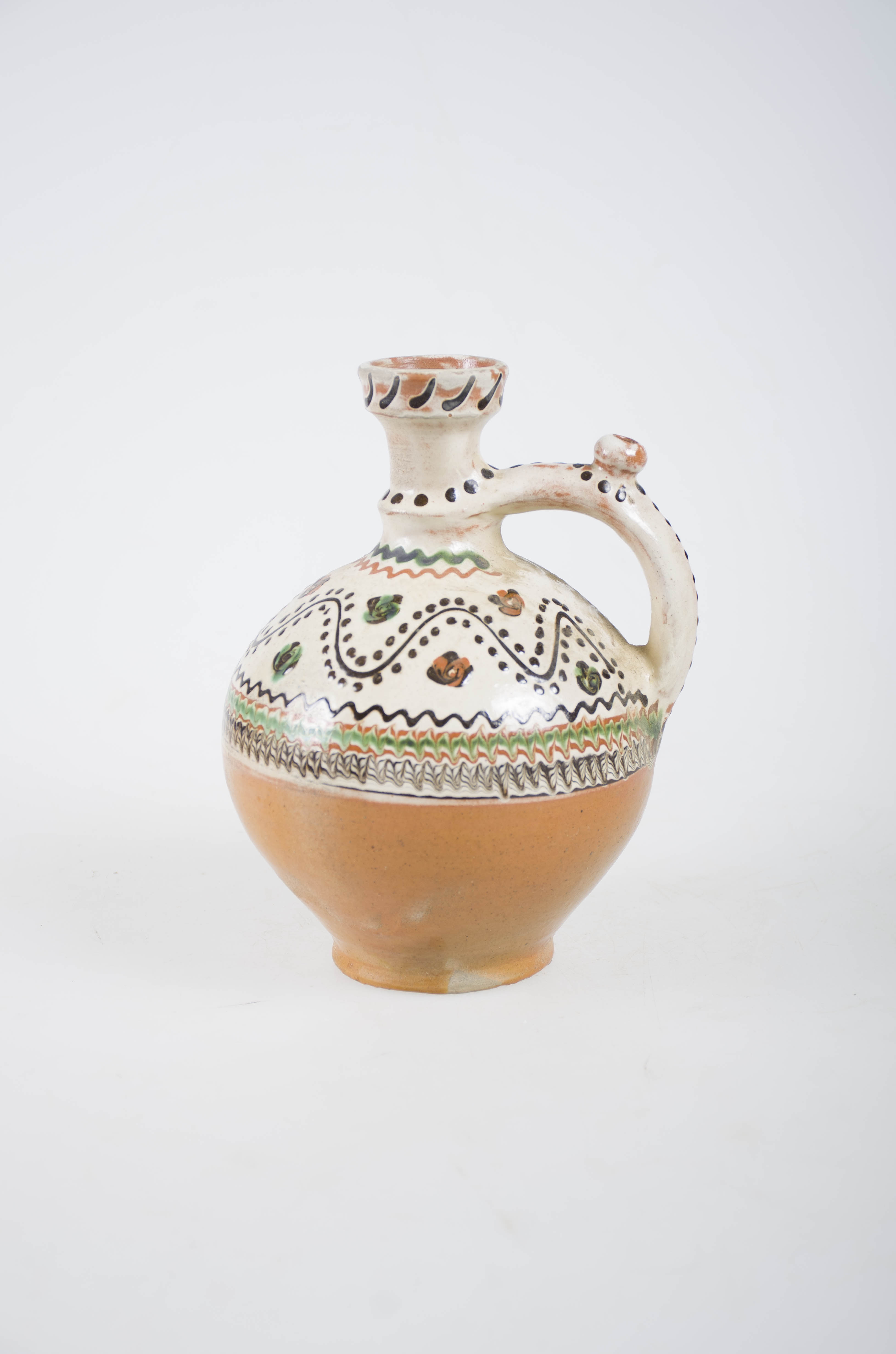 Vas ceramic sferic, ce prezintă inel de susținere din care pornesc pereții arcuiți puternic și care în partea superioară se îngustează terminându-se cu un gât înalt având un mic inel de susținere, buza dreaptă. De la inelul de susținere se prinde mănușa până la diametrul maxim. Mănușa prevăzută cu "țâță". Ornamentul realizat cu cornul este dispus pe marginea superioară care este angobată și care se compune dintr-o porțiune jirăvită, o linie ondulată cu puncte de o parte și de alta și cu "boboci" din loc în loc. La baza gâtului două linii ondulate, mănușa cu puncte, iar marginea buzei cu linii. Jumătatea inferioară are culoarea lutului ars. Smălțuit în interior și exterior. Cromatică: fond: alb; motive: verde deschis; verde închis; cărămiziu; maro închis, Pe fundul vasului apare numele meșterului și localitatea "Ogrezeanu Stelian - Horezu".