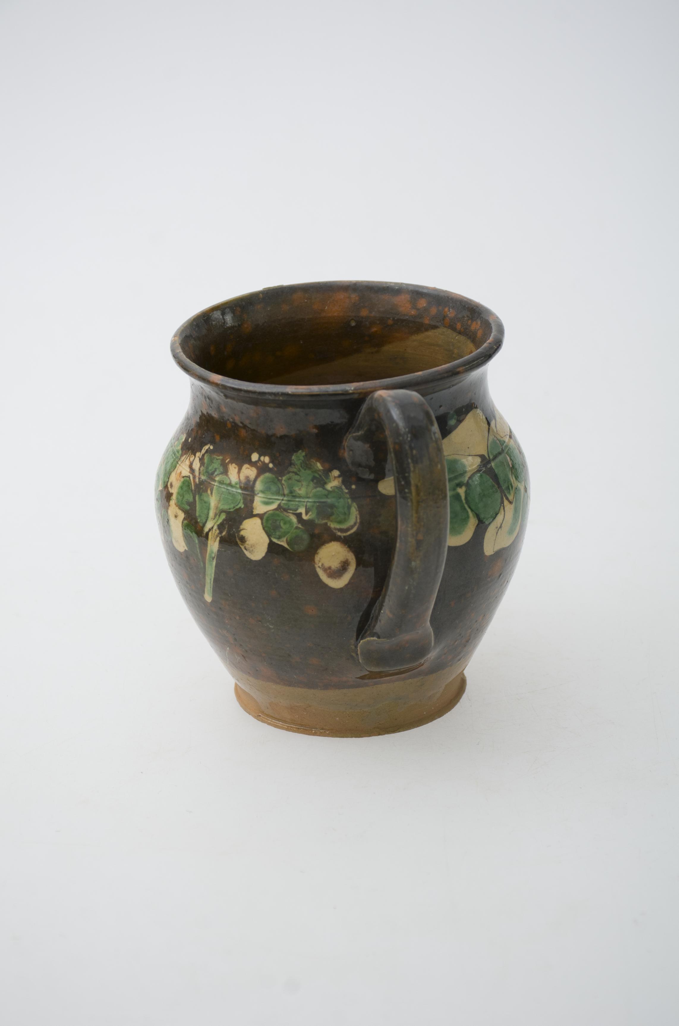 Vas ceramic de mici dimesiuni de formă bitronconică, cu baza plată, gâtul înalt și buza ușor evazată. Cană prezintă o toartă care pornește din zona buzei, și se unește cu vasul în zona diametrului maxim al acestuia. Peste stratul de angobă maro închis, în partea superioară a fost realizat decorul, compus dintr-un registru pictural dispus concentric, compus din pere de culoare verde și maro galben. În partea inferioră, în proximitatea fundului, vasul prezintă cromatică maro dechis, nefiind pictat sau smălțuit. Cromatică: fond: maro închis, maro deschis; motive: verde, galben.