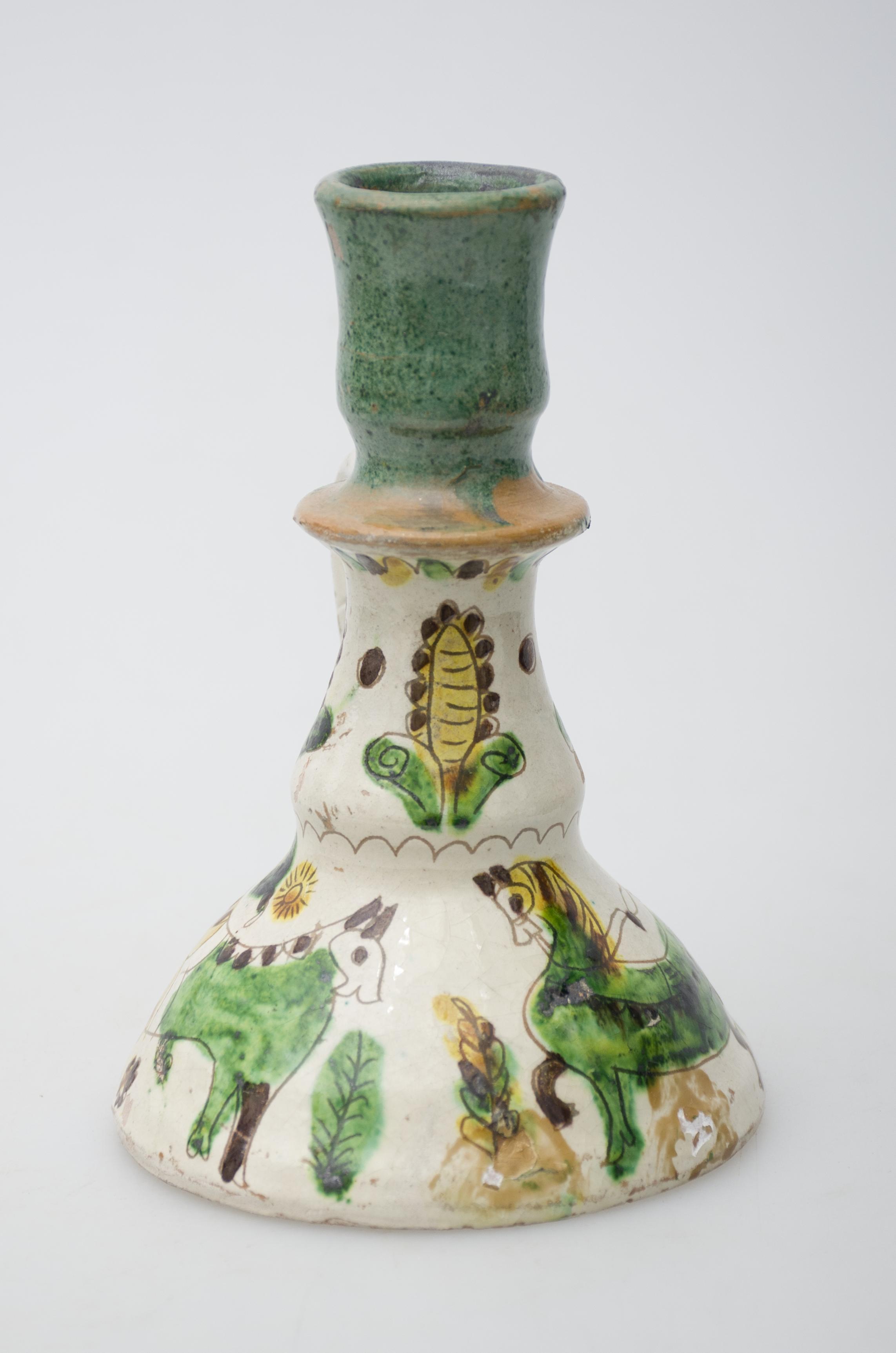 Obiect ceramic de uz casnic, format dintr-un picior tronconic, din care pornește un braț în care se introduc lumânările. Sfeșnicul prezintă o toartă, care pornește din zona gâtului și se unește cu obiectul în zona evazării piciorului. Decorul pictural, realizat pe toată suprafața obiectului, este compus din motive fitomorfe, geometrice, zoomorfe și antropomorfe. Primul registru, situat pe baza evazată, este format dintr-un cal care trage după el un atelaj în care se află un trâmbițaș. În partea opusă un animal fantasmagoric. Registrul superior este compus din trei flori cu pistilul pronunțat, și despărțite de câte un punct. Gâtul și buza vasului au fost decorate cu culoarea verde. Cromatică: fond: alb; motive: verde, maro închis, galben, negru.
