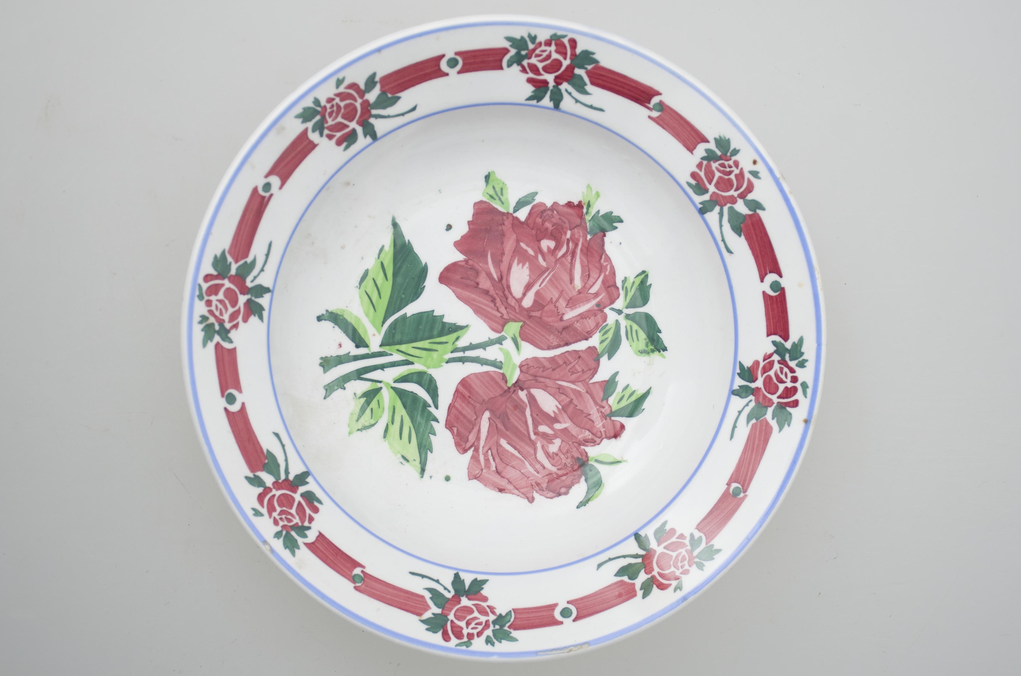 Farfurie din porțelan, de uz casnic, întrebuințată în gospodărie la decorarea pereților casei. Formă circulară, adâncită. În interioriul vasului se identifică un decor un fitomorf, motive vegetale și florale și un decor geometric. Pe margine este decorat cu motive florale, despărțite de motive geometrice: linie, punct, care sunt încadrate de două cercuri concentrice albastre; motive florale în centru (trandafiri). Pe fund prezintă găuri pentru agățare, trei cercuri concentrice și o ștanță cu o inscripție în limba german, precum și o emblemă/blazon. Cromatică: fond alb; motive: roșu, verde, albastru.