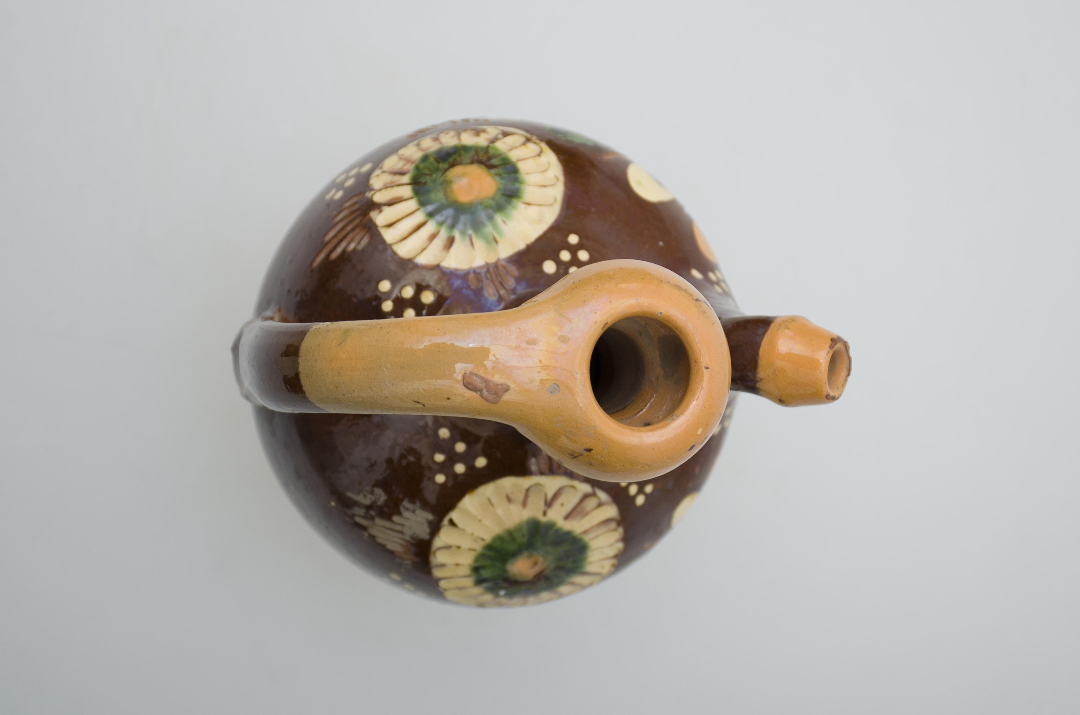 Recipient ceramic de formă sferoidală cu gâtul lung cu manșon, și buza rotundă. Smălțuit până aproape de fundul vasului. Prezintă o toartă lată și arcuită ce pleacă de la baza gurii, spre partea mediană a vasului. Din umerii ibricului, opus toartei, pleacă o ”țâță” arcuită, pentru scurgerea lichidului. Intens stilizată cu motive geometrice (puncte, linii dispuse vertical) și vegetal-florale (crenguțe, flori cu petale multiple), realizate cu pensula prin pictare. Cromatică: fond: maro; cărămiziu; motive: alb, verde, crem.