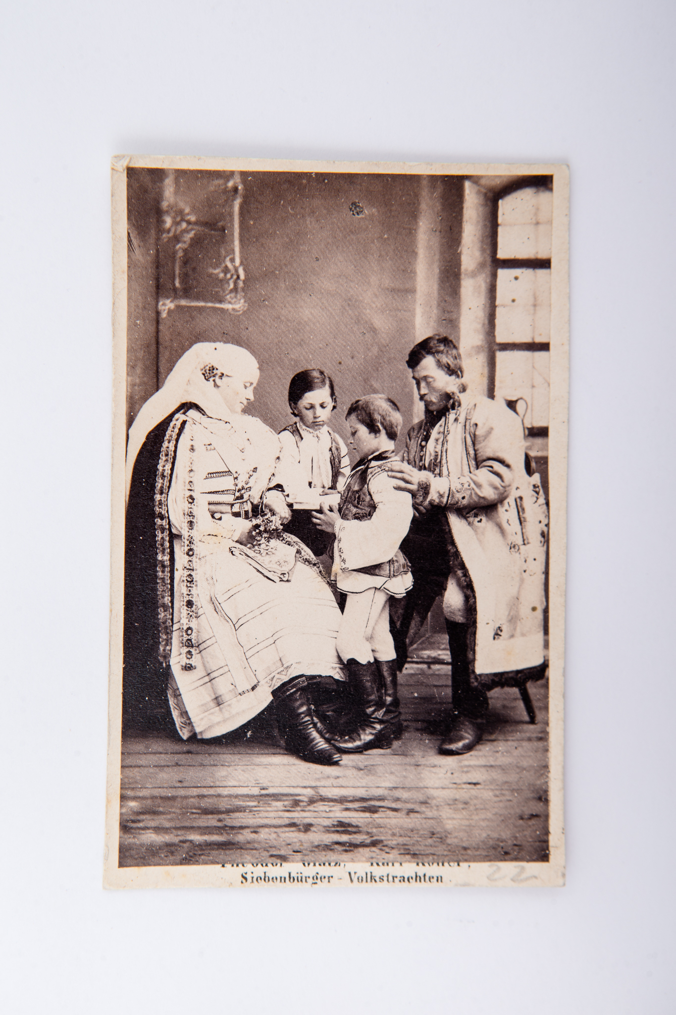 Fotografie de atelier cu decor pictat și elemente de recuzită: masă, scaune, imprimată direct (POP - printing out process) pe hârtie albuminată, reprezentând o familie de sași așezați la masă. Cei doi părinți sunt așezați pe scaune, iar copiii stau între cei doi. Fotografia format Carte de Visite este lipită pe un carton gros, crem. Pe carton numele atelierului fotografic și text imprimat, parțial vizibil, în limba germană: „Theodor Galz, Karl Koller / Siebenbürger - Volkstrachten” (Theodor Galz, Karl Koller / Costum popular transilvănean) V: Pe verso însemnare manuscrisă: „Bistrița”, MCPR 807.