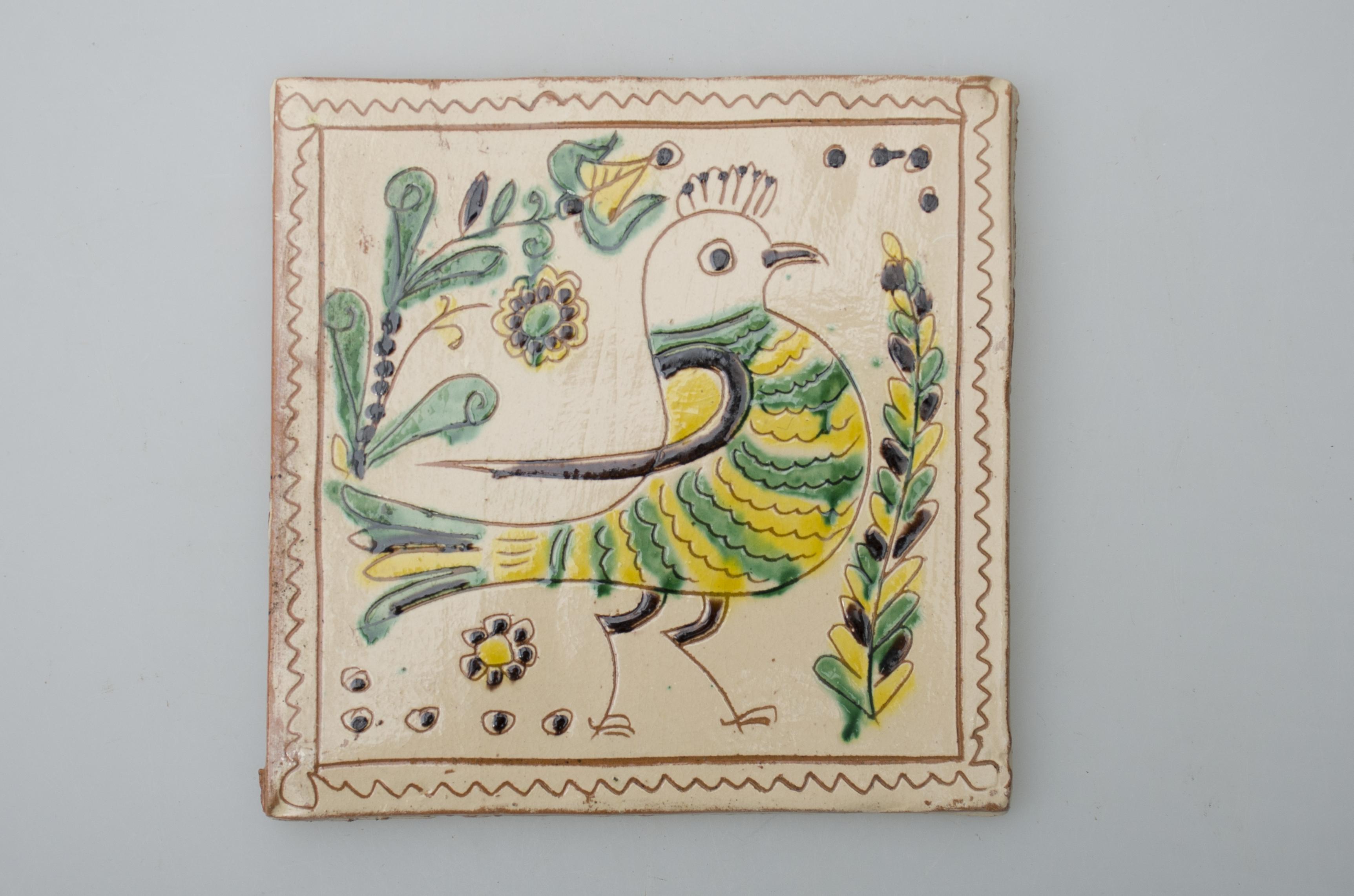 Piesă ceramică de formă pătrată, smălțuită și angobată, cu rol decorativ. Laturile pătratului sunt marcate de două chenare, primul vălurit, al doilea în linie dreaptă. Ornamentată cu motive avimorfe, geometrice și floral-vegetale: o pasăre mare (o pupăză?), dispusă central, care privește spre dreapta, fiind încadrată în patru flori diferite. Pe două din margini sunt pictate puncte negre în cercuri maro. Motivele au fost la început incizate, iar apoi pictate. Pe spate, cahla este prevăzută cu un orificiu pentru prinderea aței. Tot aici, este inscripționat numele meșterului olar, "Colibaba". Cromatică: fond: alb; motive: verde, galben, maro, negru.