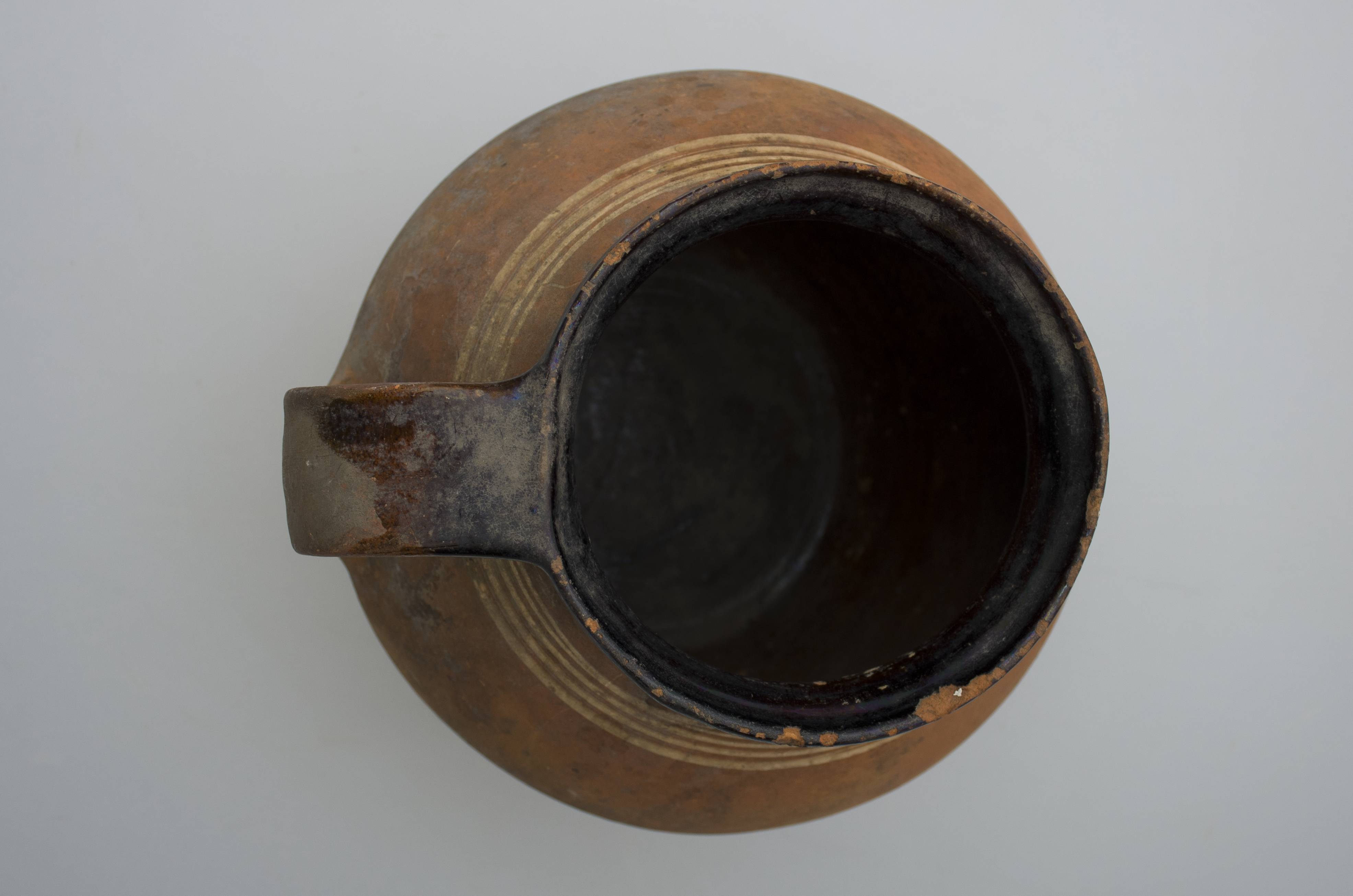 Vas ceramic smălțuit de formă bitronconică, cu baza plată, pântecul bombat și buza profilată. Oala prezintă o toartă care pornește de la buză și se unește cu vasul în zona diametrului maxim al acestuia. Pe „umerii” recipientului a fost realizat decorul geometrizant: patru linii circulare succesive de vopsea albă. Interiorul oalei, buza și partea superioră a toartei prezintă un strat maro închis de angobă peste care a fost un strat gros de smalț. La exterior, vasul nu a fost smălțuit. Oala era folosită în gospodărie pentru prepararea termică a alimentelor. Prezintă uzură funcțională. Cromatică: fond: cărămiziu, maro închis; motive: alb.