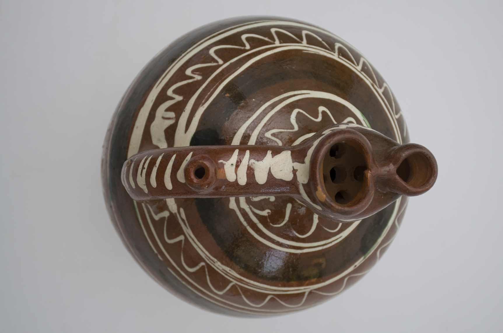 Vas ceramic smălțuit de formă ovoidală, bombat în partea mediană, cu gât strâmt terminat cu o gură bilobată.cu ciur. Prezintă o toartă puternic arcuită cu „țâță'' și piatră sunătoare în interior. Decorații reprezentând trei linii albe vălurite dispuse circular încadrate de linii drepte concentrice. Mijlocul gâtului și toarta sunt conturtate cu câte o linie ondulată. Pete verzui dispuse aleatoriu pe întreg corpul vasului. Cromatică: fond: brun; motive: alb; verde.