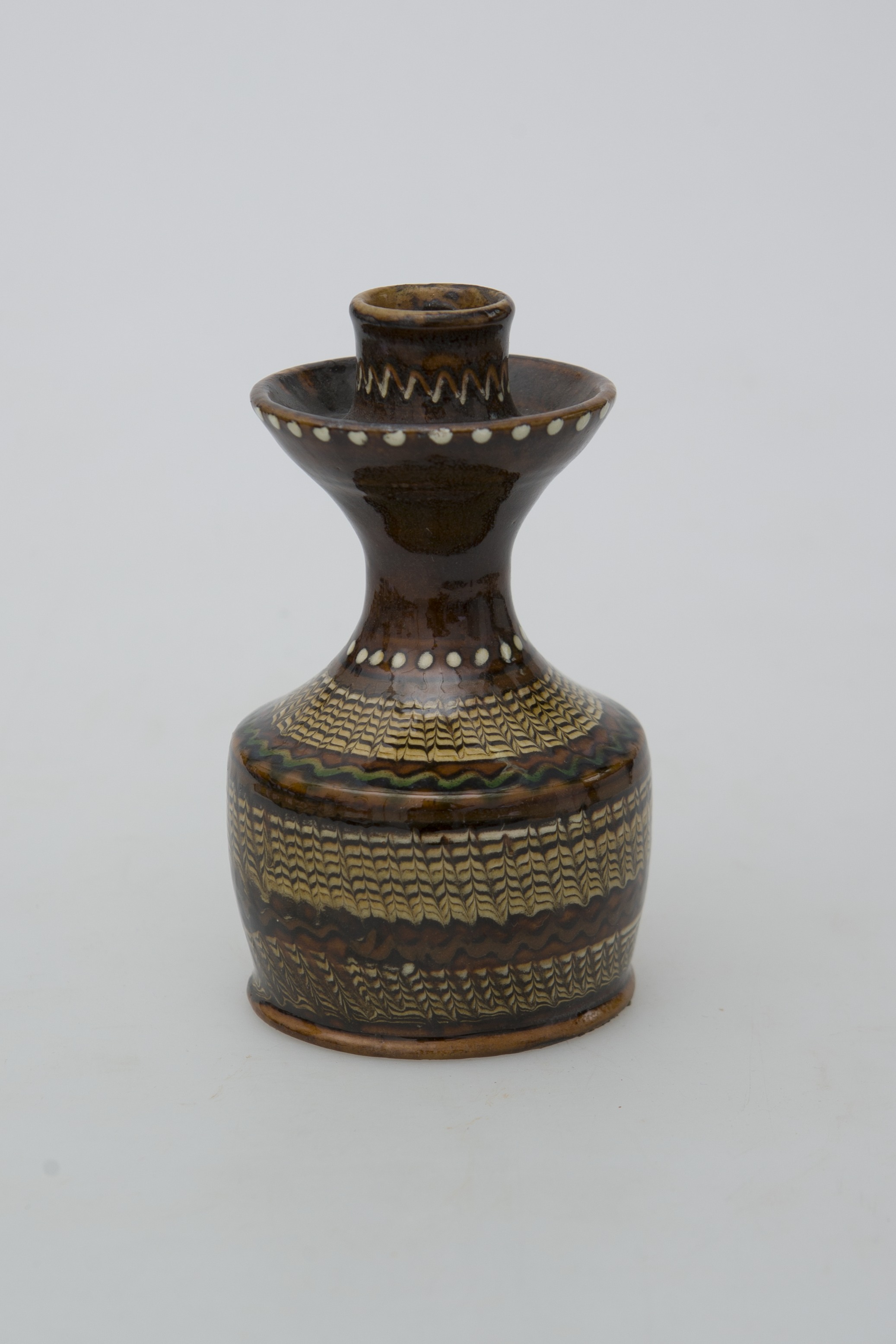 Recipient ceramic tubular, folosit ca suport de lumânare. Baza, fără fund, foarte înaltă cu pereții drepți ce se continuă printr-un gât îngust, înalt, lățindu-se în partea superioară pe marginea care se evazează la exterior. Este prevăzut un șanț pentru ținerea cerii topite. Partea superioară prevăzută cu un orificiu înalt pentru fixat lumânarea. Decorații jirăvite pe întreg corpul, linie verde ondulată pe umăr, linie punctiformă circulară la baza gâtului și pe inelul reliefat, și o linie în zig-zag în partea superioară a gâtului. Smălțuit în exterior. Cromatică: fond: maro închis; motive: alb; verde; maro; negru; bej.