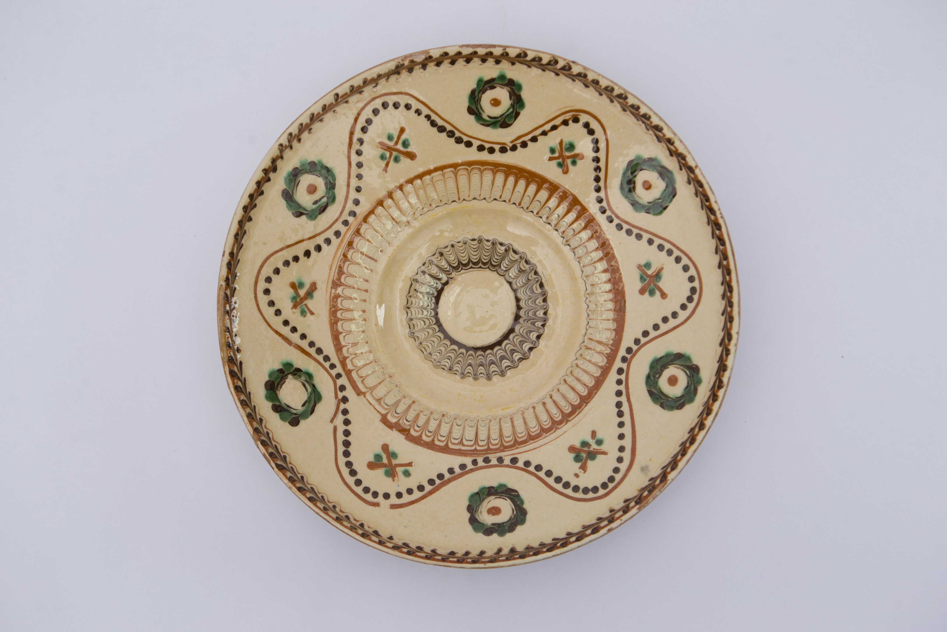 Vas ceramic de formă discoidală cu baza plată a vasului din care se arcuiesc pereții; marginea farfuriei este evazată spre exterior cu buza rotunjită. Ornamentul realizat cu cornul este dispus pe toată suprafața în registre structurate concentric. Fundul farfuriei are un cerc jirăvit urmează un ornament de arcuri la fel închise într-un cerc și o linie ondulată cu puncte, dublată de o altă linie continuă ondulată, în ultimul registru. Între ondulațiile liniei ondulate continue se găsesc ”cununi'' închise, în verde și nergru, cu o bulină maron în mijloc; iar între ondulațiile liniei liniei punctiforme ”X''-uri însoțite de puncte. La buza vasului dispusă o linie circulară. Smălțuit în interior; exteriorul culoarea lutului ars. La posterior baza inelară este perforată pentru atârnare. Cromatică: fond: crem; motive: maro închis; maro roșcat; verde; alb.