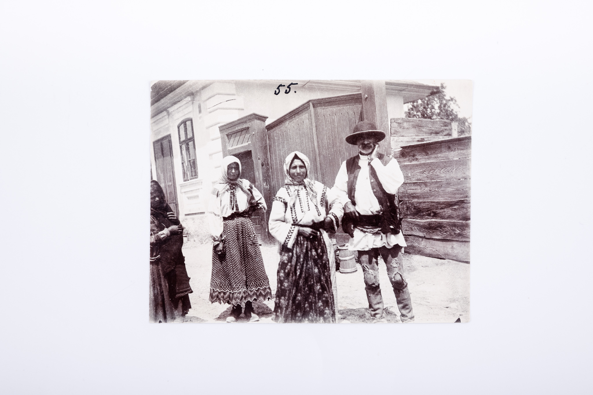 Fotografie de exterior realizată pe o uliță, lângă poarta de intrare într-o gospodărie. Prezintă trei femei și un bărbat de etnie rromă, în port specific. Fotografia este imprimată pe hârtie cu gelatino-bromură de argint, după un clișeu pe sticlă. În partea de sus, însemnare manuscrisă: „55.” V: Însemnare manuscrisă, cu creionul, în limba germană: „Zigeuner / Dörfer” (Țigani de la sat). Număr de Inventar MCPR 2304.