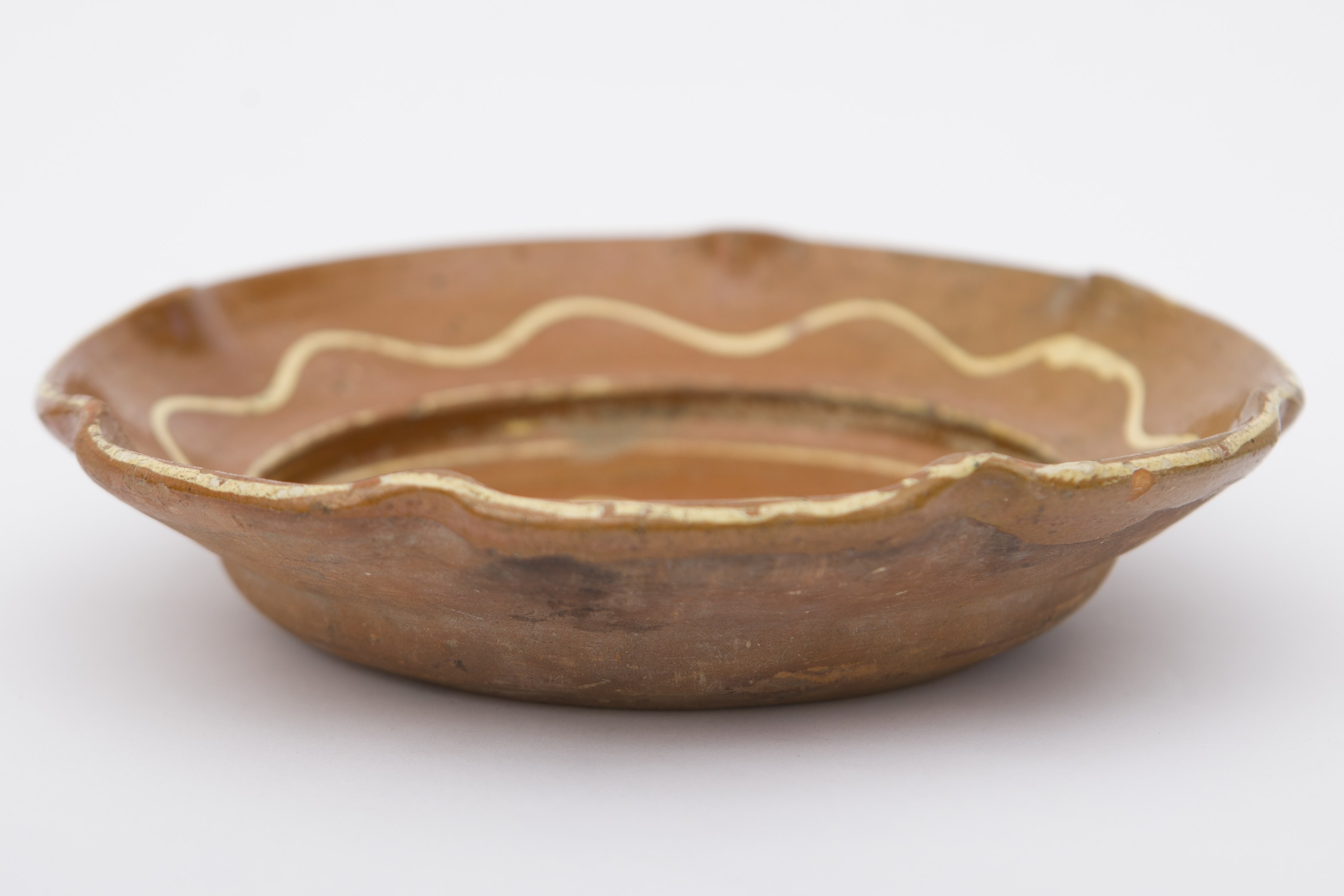 Piesă ceramică, smălțuită în interior, de formă tronconică cu baza plată, pereții înalți și buza vălurită. Peste stratul maro deschis de angobă au fost realizate prin pictare decorații fitomorfe, ilustrând o floare cu petale albe și frunze maro închis, dispusă central, pe fundul vasului, fiind încercuită de tulpina sub forma unei linii spiralate. O altă linie circulară, însă în valuri, ornamentează buza vasului, formând astfel o a doua floare. Farfuria era folosită pentru servirea hranei sau ornamentarea interioarelor. Prezintă uzură funcțională. Cromatică: fond: cărămiziu (exterior); maro deschis (interior); motive: alb; maro închis.
