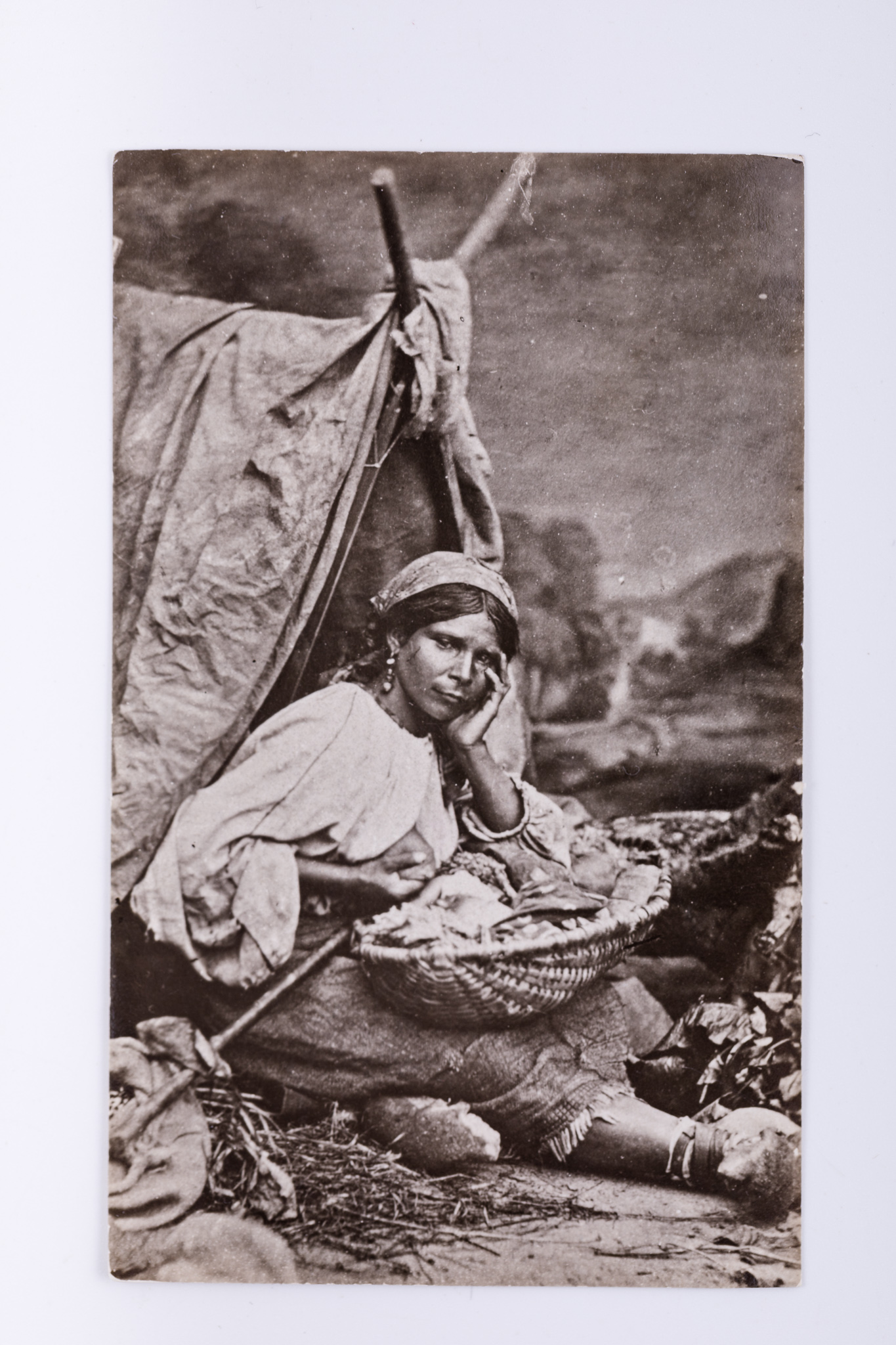 Fotografie de atelier cu elemente de recuzită: cort, elemente vegetale, merindeață. Imaginea prezintă o femeie de etnie rromă, în port specific. Aceasta stă așezată, ține în poală un coș împletit în care se află un copil pe care îl alăptează. Fotografia este o reproducere pe hârtie cu gelatino-bromură de argint, replică din secolul al XX-lea după o fotografie realizată după un clișeu pe sticlă. V: Însemnare manuscrisă, cu creionul, în limba germană: „Volkstypen 21 / K.I.2 / Zigeuner mit Kind / Tipuri etnice”. Ștampilă: „Baron Brukenthal`sches Museum in Hermannstadt”. Număr de Inventar MCPR 2298.