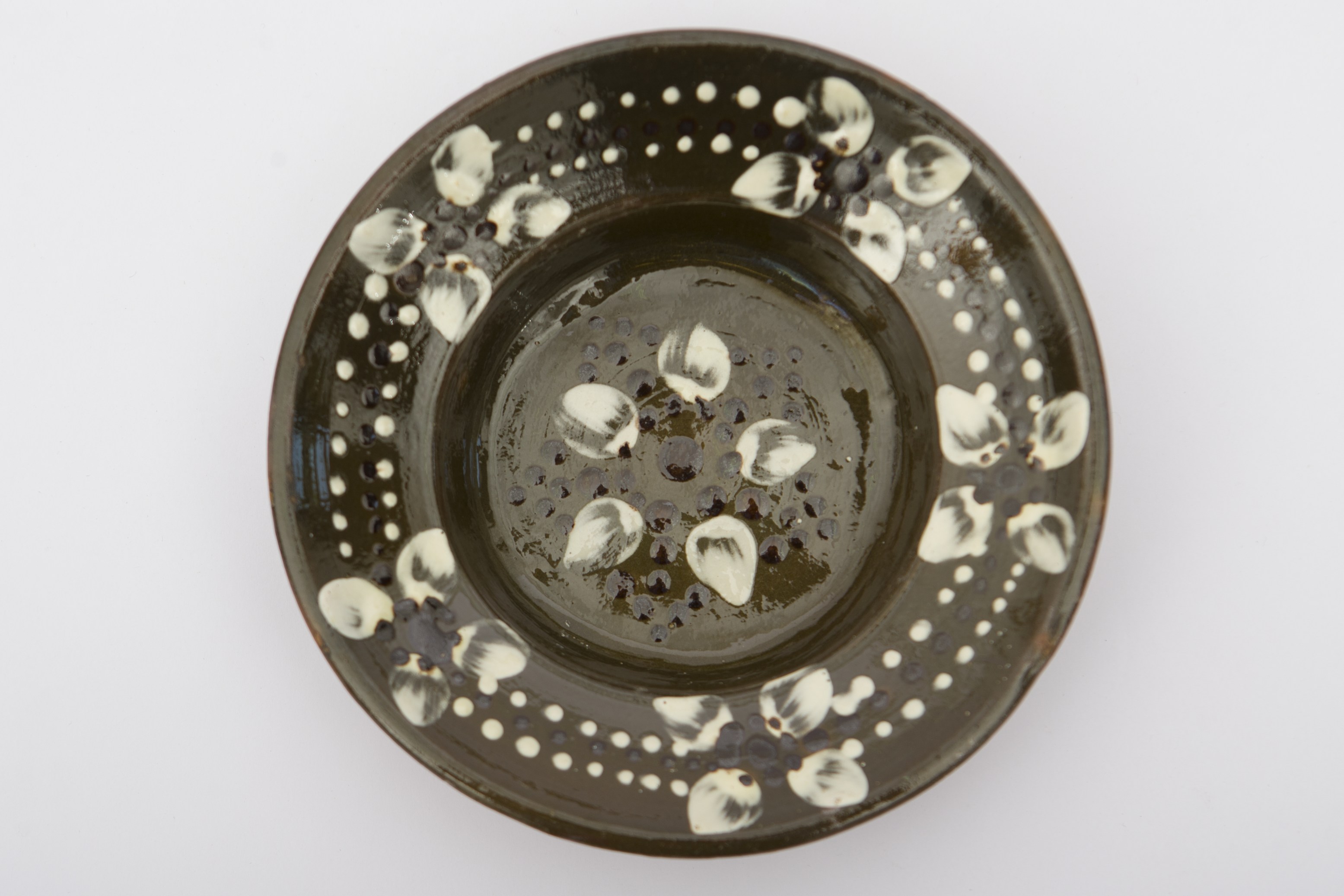 Piesă ceramică smălțuită în interior, de formă tronconică cu fundul plat, marginile ușor ridicate și buza dreaptă. Peste stratul verde închis de angobă a fost realizat decorul fitomorf și geometrizant stilizat, compus din: șase flori, una dintre ele având cinci petale albe și un punct negru în mijloc, încadrată de puncte negre care crează o stea cu cinci colțuri. Celelalte flori se află pictate pe gura vasului la o distanță egală una față de cealaltă. Florile cu câte patru petale albe cu un punct negru în mijloc, sunt unite de trei șiruri de puncte dispuse circular, șirul din mijloc având puncte de culoarea neagră, celelalte în alb. Recipientul dispune în partea din spate de o tortiță de mici dimensiuni pentru suspendare de perete. Blidul era folosit în gospodărie pentru decorarea interioarelor. Prezintă uzură funcțională. Cromatică: fond: cărămiziu (exterior); verde închis (interior); motive: alb; negru.