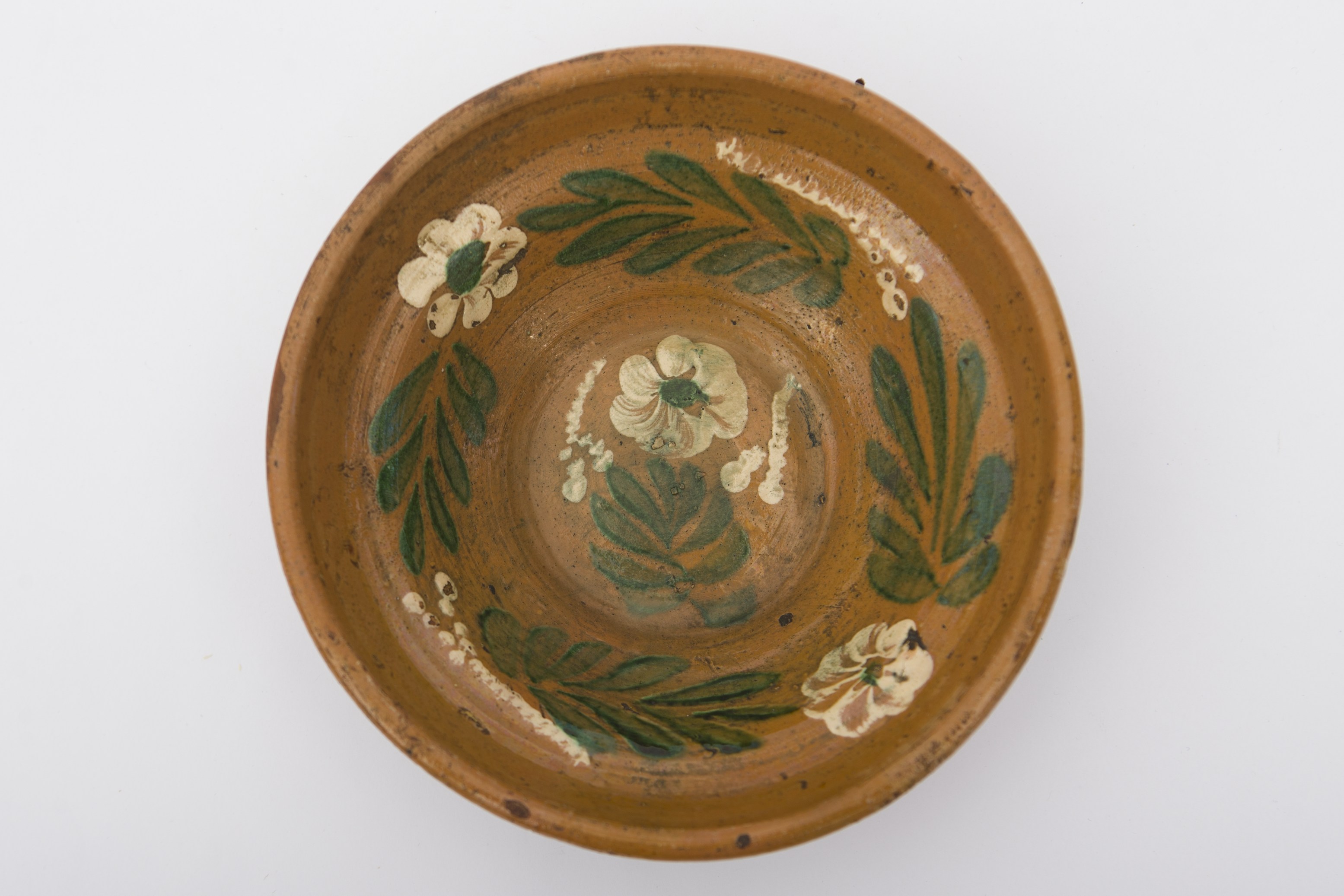 Vas ceramic, smălțuit în interior, de formă tronconică cu baza plată, pereții ridicați și buza profilată. Peste fond maro deschis decor fitomorf realizat prin pictare cu pensula, ilustrând trei flori cu petale albe, cu câte un punct verde în mijloc. Una dintre flori se află pe fundul vasului, având la bază frunze verzi și fiind încadrată de două motive punctate. Celelalte două flori se află dispuse pe pereții interiori ai vasului în alternanță cu frunze verzi și motive punctate. Pe spate prezintă o tortiță de mici dimensiuni pentru suspendare. Blidul era utilizat în gospodărie pentru servirea hranei. Prezintă uzură funcțională. Cromatică: fond: cărămiziu (exterior); maro deschis (interior); motive: verde; alb.