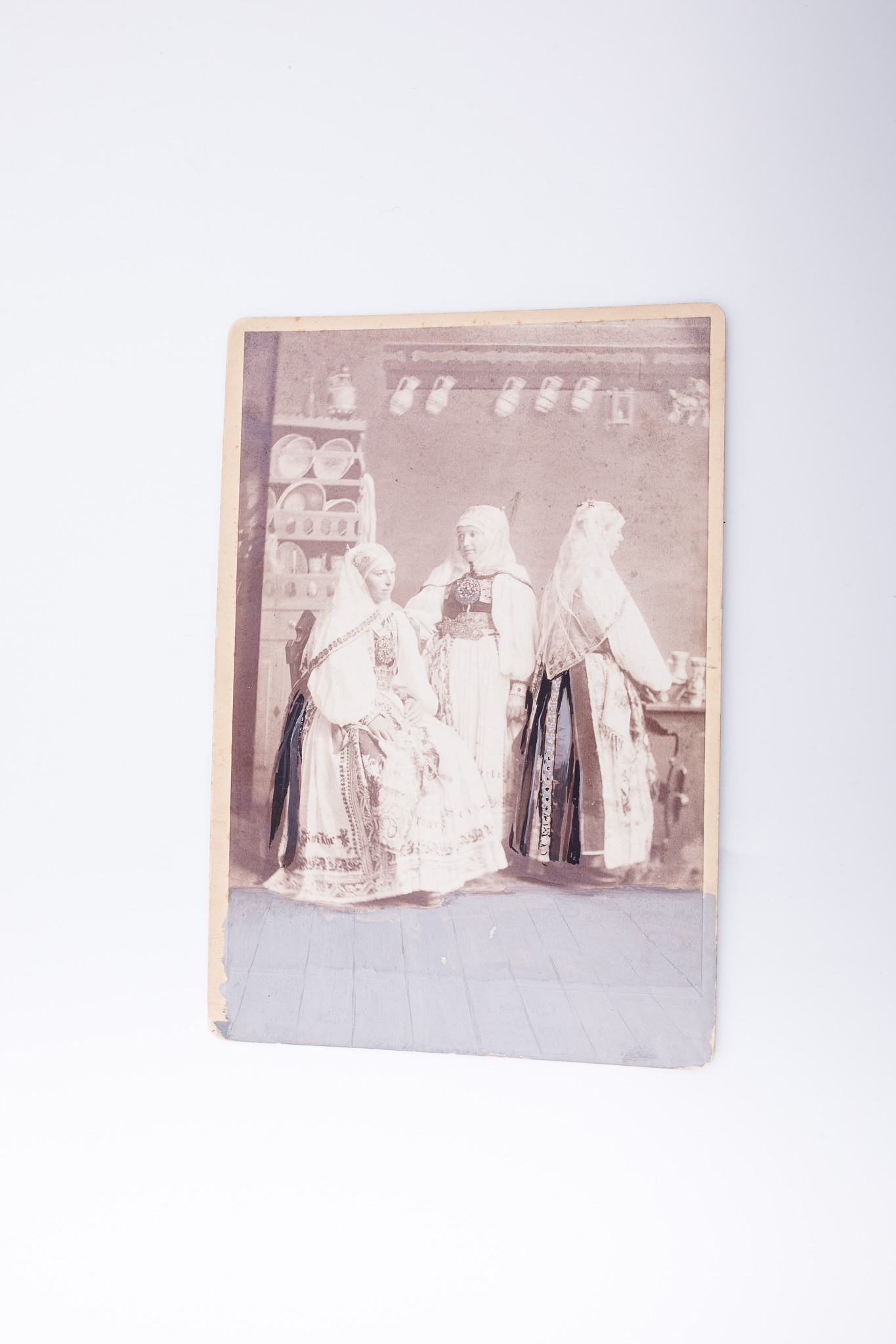 Fotografie de atelier, cu decor pictat și elemente de recuzită (masă, ladă pictată, ceramică). Prezintă trei tinere în port popular săsesc de sărbătoare din Daia, județul Sibiu. Fotografia este lipită pe un carton de culoare crem. Anumite părți ale costumului sunt evidențiate cu negru și gri, iar podeaua este colorată cu culoare gri dincolo de marginile hârtiei sensibilizate, pe toată parte de jos a cartonului. V: Ștampile: „Emil Sigerus / Hermannstadt”, „Direktor Emil Sigerus / Hermannstadt”. Însemnare manuscrisă, cu creionul în limba română și germană: „Daia / Jos. Drotleff / Hermannstadt”. Putem presupune că fotografia a fost ulterior tipărită de către tipografia Jos. Drotleff, din Sibiu. Marca atelierului fotografic, cu zece medalii și text de reclamă în limba germană: „No. 71 / Atelier / Kamilla Asboth / vormals / Th. Glatz`s erben / Hermannstadt / Vervielfältigung vorbehalten / Die Platten werden zu Nachbestellungen aufbewahrt” („Nr. 71 / Atelier / Kamilla Asboth / Moștenitoarea lui Th. Glatz / Sibiu / Reproducere rezervată / Plăcile sunt păstrate pentru comenzi ulterioare”); număr de Inventar MCPR 2509.
