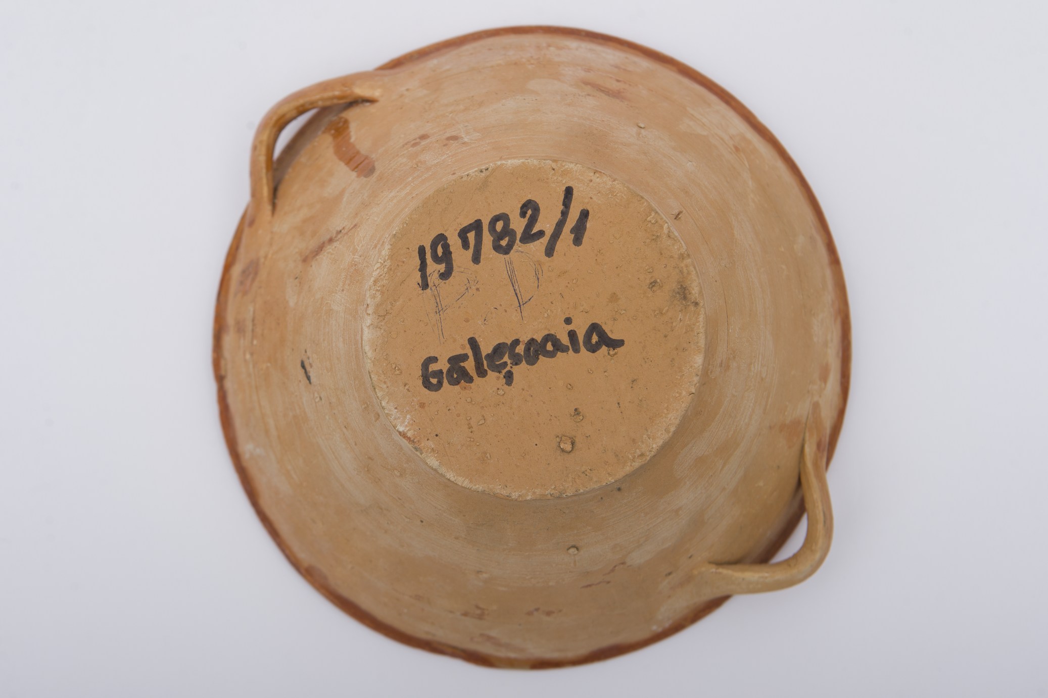 Vas ceramic, smălțuit în interior, de formă rotundă, profil tronconic și pereți evazați. Sub buză prezintă două tortițe laterale pentru manipulare. Pe fundul vasului amplasată o ,,crenguță de brad', sugerată de o linie dreaptă de unde se desprind latelar linii diagonale dispuse descrescător spre vârf, înscrisă într-un cerc concentric. Încă două asemenea cercuri fiind dispuse pe pereții interiori, împreună cu un șir de scurte linii albe și negre palmate dispuse circular. Buza dreaptă conturată cu o linie ondulată dispusă circular. Cromatica: fond: cărămiziu; motive: alb, negru.