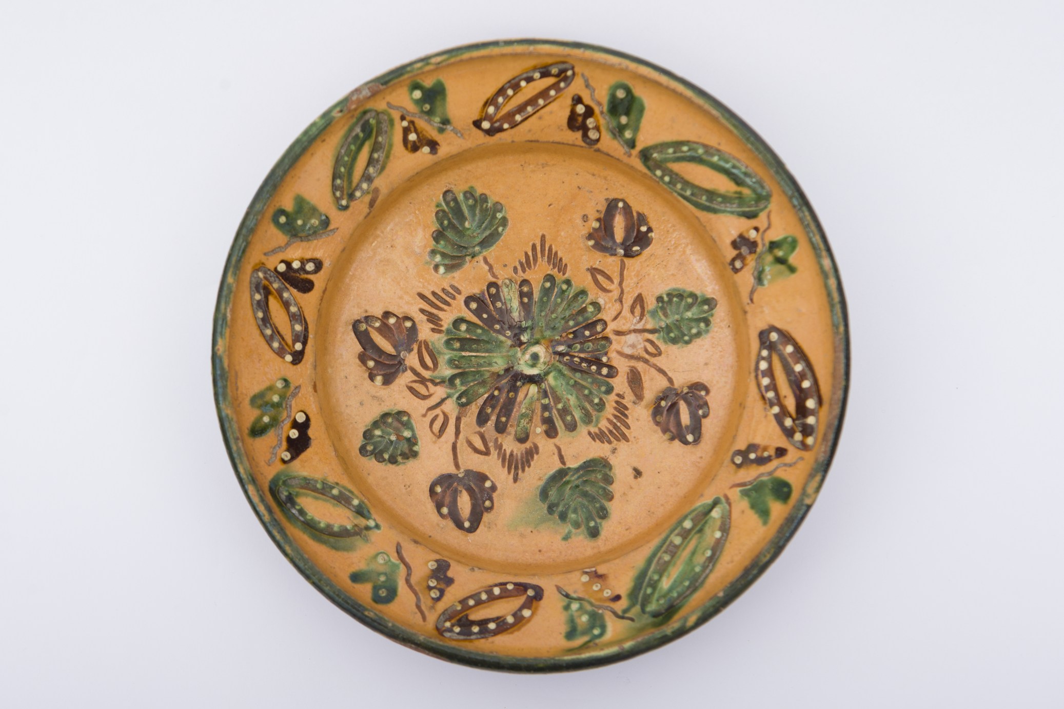 Vas ceramic cu fund plat, smălțuit în interior, cu margini ușor răsfrânte în interior. Buza este oblică, cu margine rotunjită. Ornamente geometrice și vegetale pe toată suprafața vasului. Peste decorul fin incizat se suprapune pictarea cu culori. Motive ilustrând rozeta centrală cu lalele împrejur lucrate cu scoaba, împreună cu alte motive floral-vegetale care se desprind din motivul central, despărțite de grupe de linii palmate scurte. Pe marginea farfuriei șir de ovale despărție de motive fitomorfe stilizate. Buza vasului este conturată cu verde. Peste decorații se suprapun și linii punctate. Cromatica: fond; gălbui; motive: verde, brun, alb.