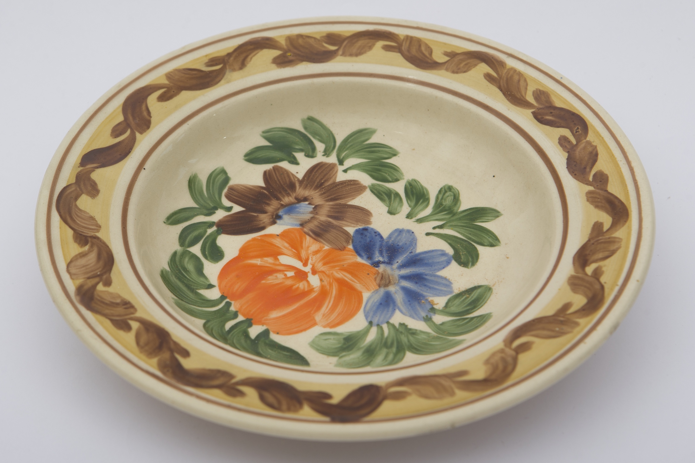 Vas ceramic smălțuit, de formă tronconică, cu baza plată, pereții ușor ridicați și buza larg evazată. Peste fondul alb au fost realizate decorații fitomorfe stilizate. Central, pe fundul vasului au fost pictate trei flori: una, mai mare portocalie și alte două mai mici, una maro cu mijlocul albastru și alta, albastră cu mijlocul maro. Întreg registrul central este mărginit de frunze verzi. Marginal, o bordură gălbuie este încadrată de două linii maro, ornamentată cu un vrej vegetal cu frunze maro, decorează buza piesei. Farfuria era folosită în gospodărie pentru servirea hranei sau decorarea interioarelor. Prezintă uzură funcțională. Cromatică: fond: alb; motive: maro; verde; portocaliu; albastru; galben.