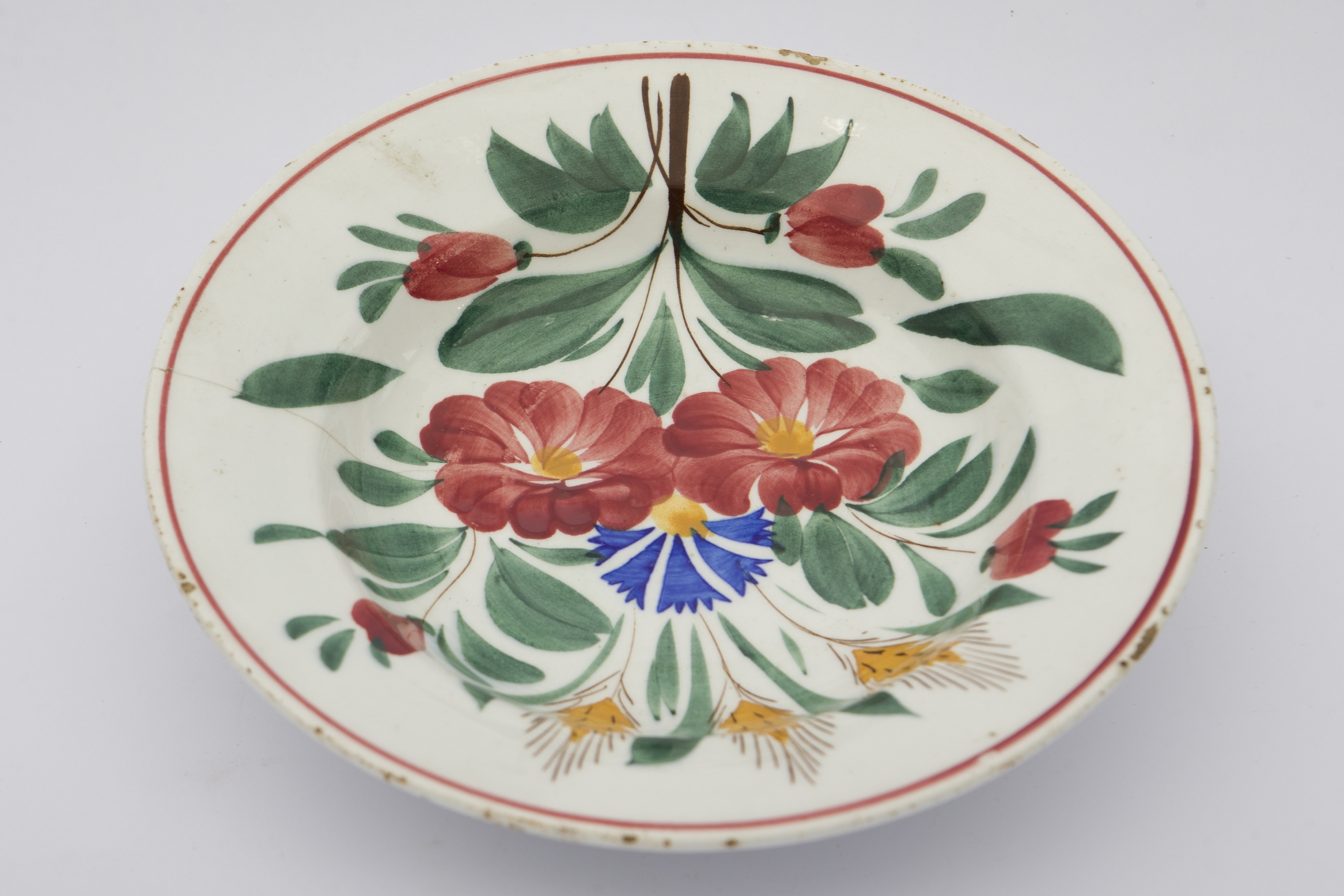 Piesă ceramică smălțuită, de formă tronconică, cu baza plată și susținută de un inel circular, pereții puțin ridicați și marginile evazate. Peste fondul alb au fost realizate decorații fitomorfe, care ocupă întreaga suprafață interioară a vasului, ilustrând trei flori, două cu petale roșii și una cu petale albastre, aflate în centrul compoziției pe fundul vasului, încadrate de frunze verzi, boboci roșii și trei spice de grâu galbene dispuse vertical, în partea de superioară a decorului. Buza vasului este conturată de o linie roșie. Farfuria era utilizată pentru servirea hranei sau decorarea interioarelor. Prezintă uzură funcțională. Cromatică: fond: alb; motive: verde; roșu; galben; albastru.