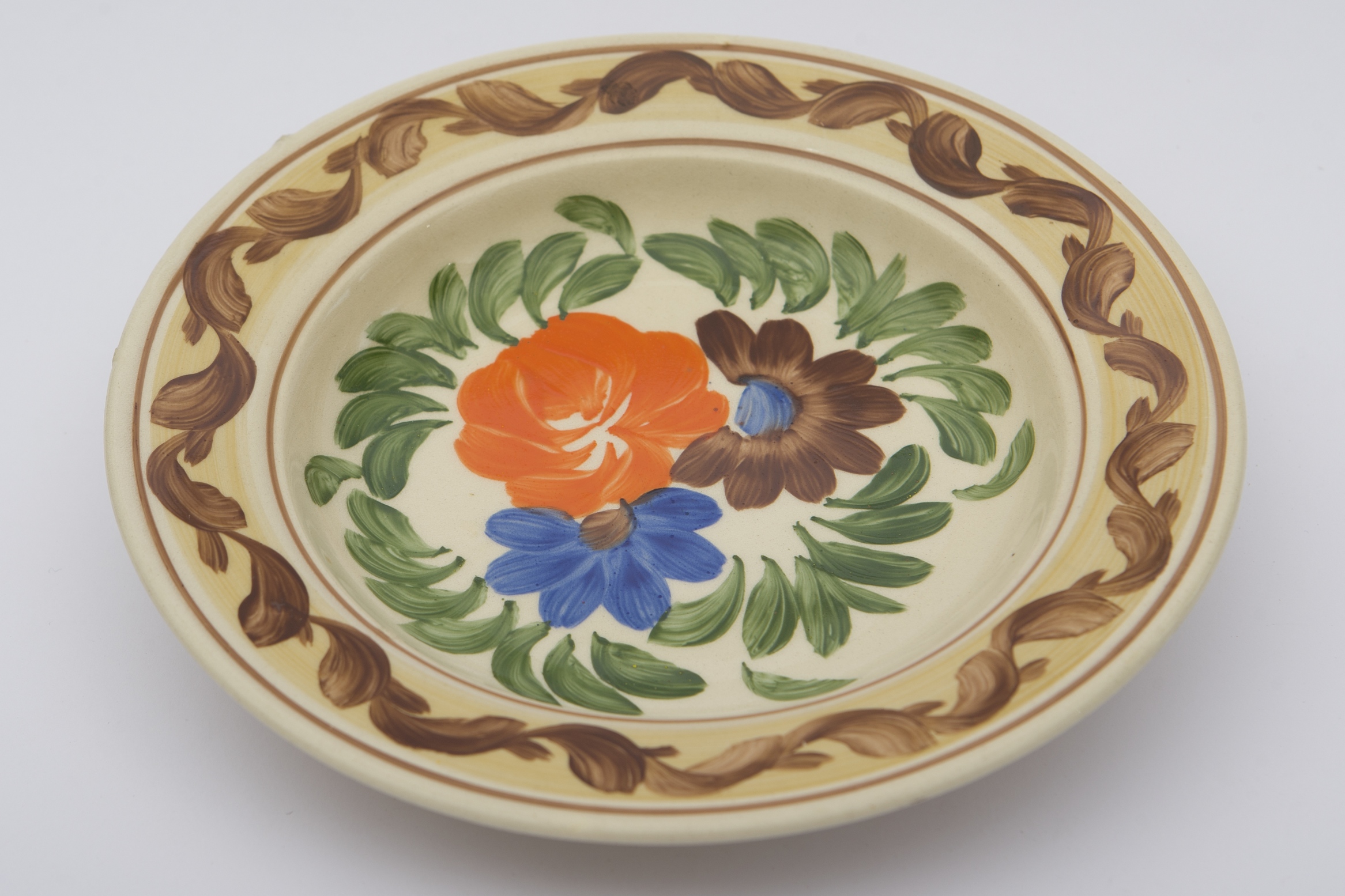 Piesă ceramică smălțuită, de formă tronconică, cu baza plată, pereții ușor înălțați și marginile evazate. Pe fond alb au fost realizate ornamentele fitomorfe stilizate. Un registru format din trei flori, prima portocalie, cea de-a doua maro, cu mijlocul albastru, ultima fiind albastră, cu mijlcul maro. Primul registru, dispus pe fundul vasului este mărginit circular de frunze verzi. Buza piesei a fost decorată cu o bordură galbuie, încadrată marginal de două linii maro, peste care a fost realizat un vrej vegetal, cu frunze maro închis. Farfuria era folosită în gospodărie pentru organizarea interioarelor, cât și pentru servirea hranei. Prezintă uzură funcțională. Cromatică: fond: alb; motive: galben; verde; portocaliu; albastru; maro.