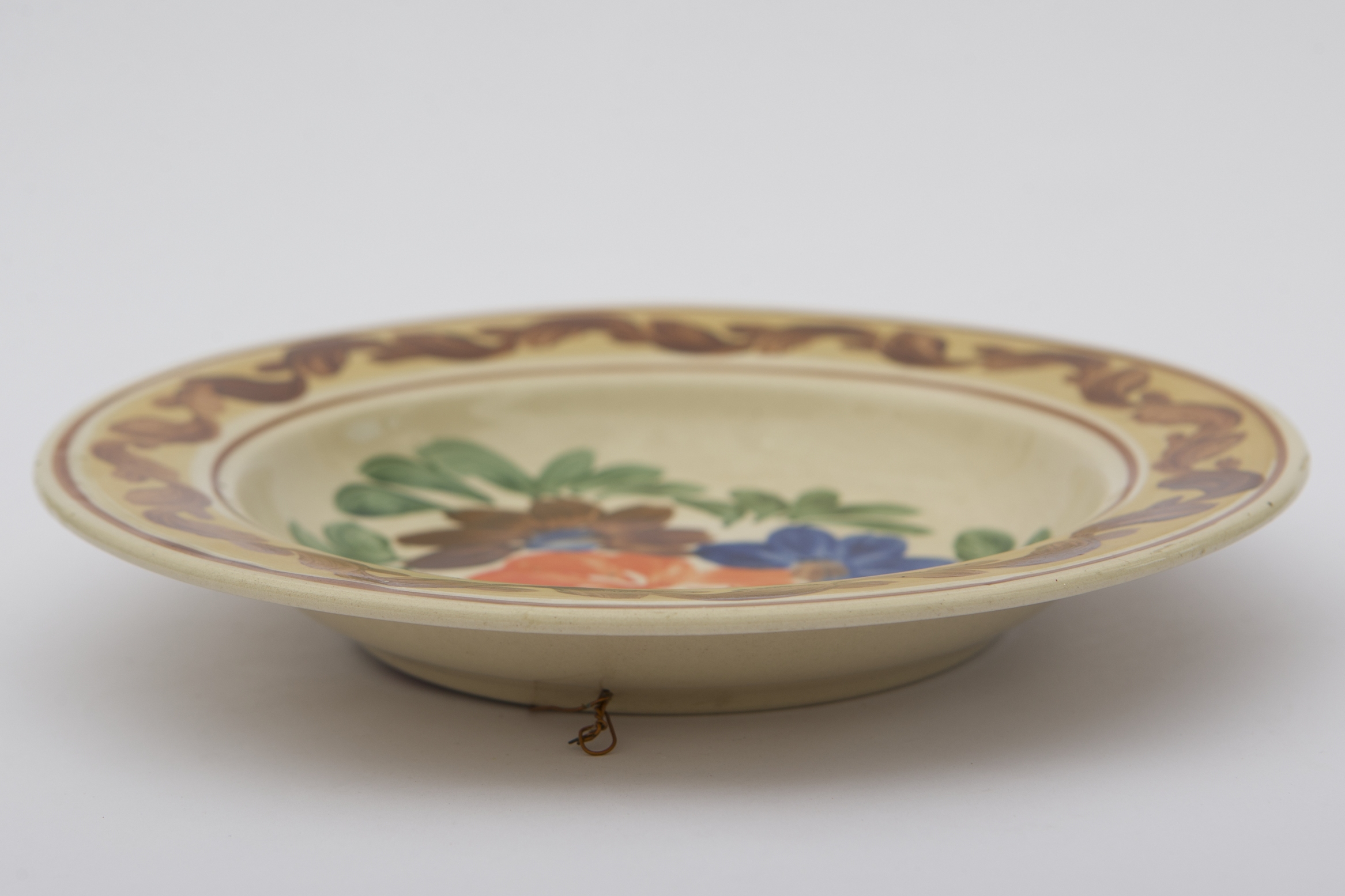 Recipient ceramic smălțuit, de formă circulară, cu baza plată susținută de un inel circular, pereții ușor înălțați și buza larg evazată. Peste fondul alb a fost pictate decorațiile fitomorfe stilizate. Central, pe fundul vasului au fost pictate trei flori: una, mai mare portocalie și alte două mai mici, una maro cu mijlocul albastru și alta, albastră cu mijlocul maro. Întreg registrul central este mărginit de frunze verzi. Marginal, o bordură gălbuie este încadrată de două linii maro, ornamentată cu un vrej vegetal cu frunze maro, decorează buza piesei. Farfuria era folosită în gospodărie pentru servirea hranei sau decorarea interioarelor. Prezintă uzură funcțională. Cromatică: fond: alb; motive: maro; verde; portocaliu; albastru; galben.