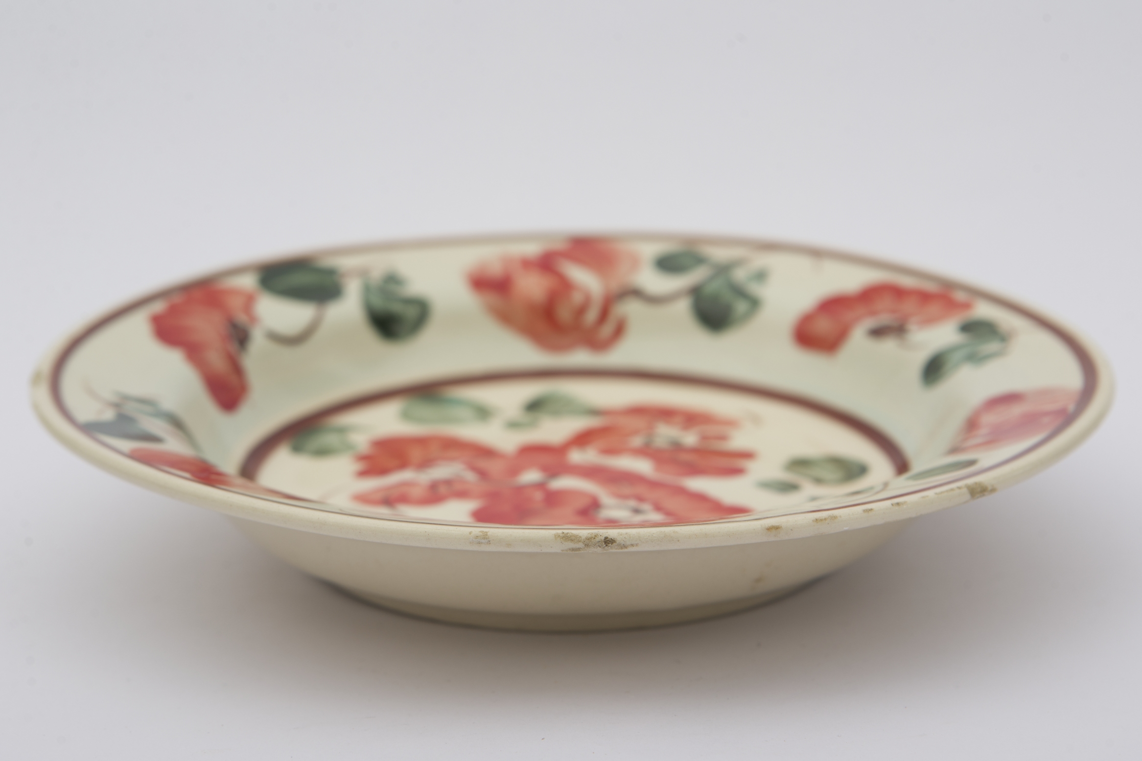 Piesă ceramică smălțuiță de dimensiuni reduse și formă cilindrică, cu baza plată și susținută de un inel circular, pereții ușor ridicați și buza larg evazată. Suprafața interioară a recipientului a fost ornamentată cu două registre decororative cu motive geometrizante și fitomorfe stilizate. Central, pe fundul vasului au fost pictați trei flori roșii cu frunze verzi, cel mai probabil maci. Marginal, pe bordura farfuriei a fost realizat cel de-al doilea registru pictural, format din șase maci cu frunze verzi, dispuși circular. Cel de-al doilea registru este încadrat de două linii circulare drepte, de culoare mov. Farfuria era folosită în gospodărie pentru decorarea interioarelor. Prezintă uzură funcțională. Cromatică: fond: alb; motive: roșu; verde; mov.