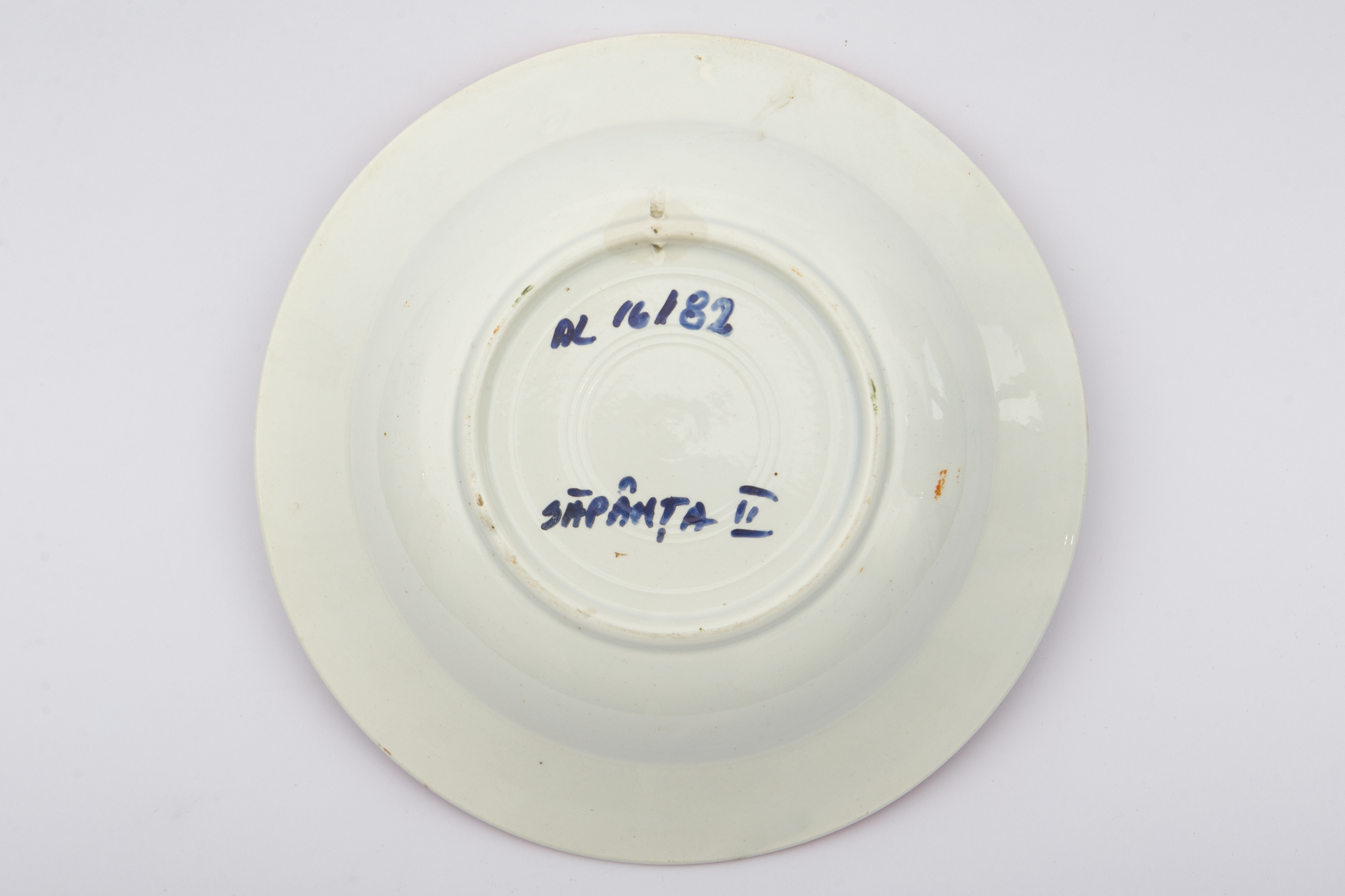 Vas ceramic de dimensiuni reduse, având formă tronconică, cu baza plată, pereții reficați și marginile evazate. Peste fondul alb au fost realizate decorațiile geometrizante și fitomorfe stilizate, dispuse în două registre. Primul registru este format dintr-un trandafir portocaliu cu frunze verzi, având în laterale câte o floare cu mijlocul albastru și petalele roșii, și câte un boboc roșu. Al doilea registru este format dintr-o tulpină circulară verde, pe care se dezvoltă trei grupuri de flori mici albastre. Alte trei grupuri, a câte trei linii portocalii oblice, însoțesc decorul fitomorf. Acest ultim registru este încadra dinspre interior de bandă groasă portocalie. Buza farfuriei a fost la rândul ei conturată cu portocaliu. Prezintă uzură funcțională. Cromatică: fond: alb; motive; portocaliu; verde; albastru.