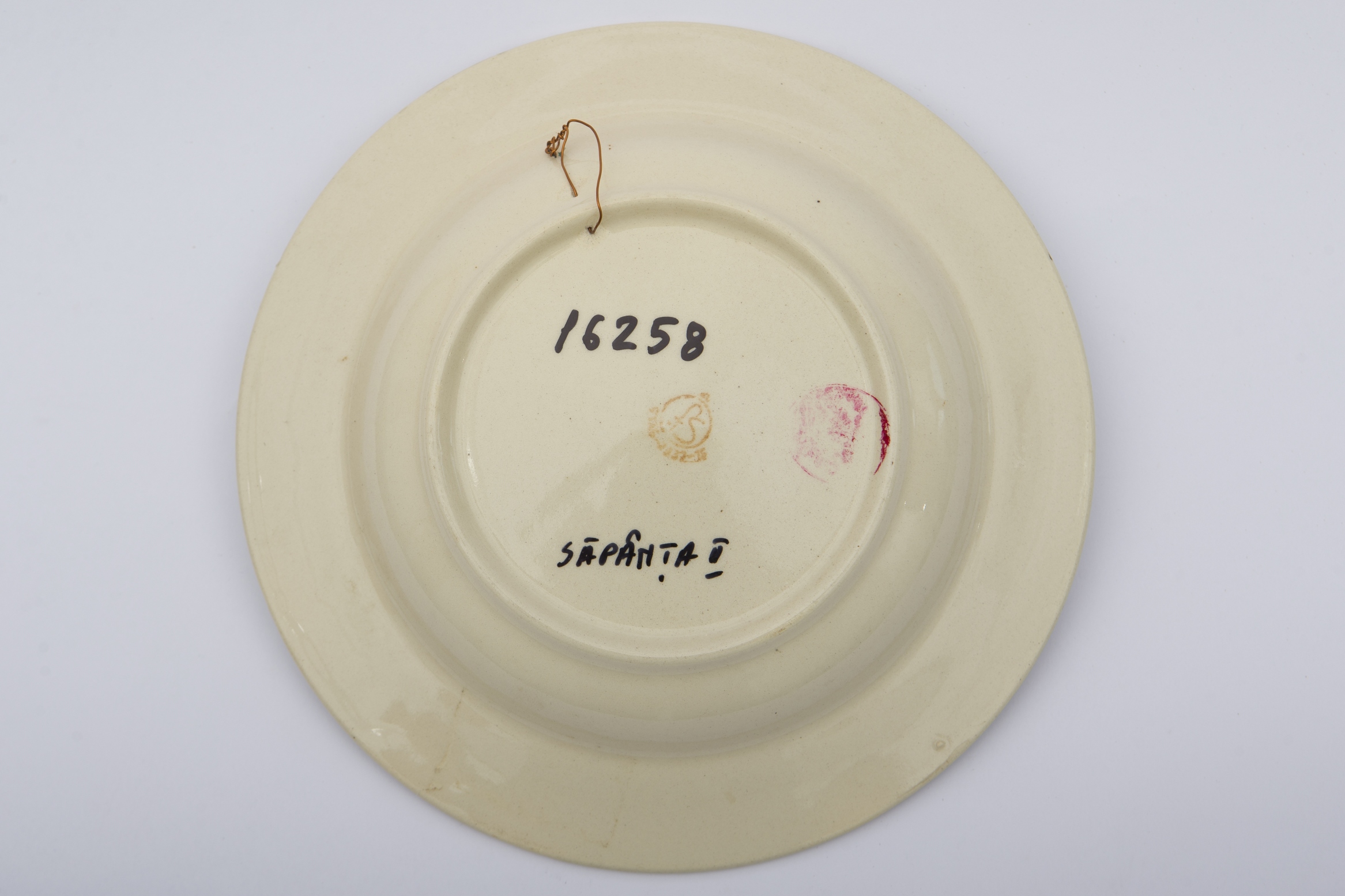Piesă ceramică smălțuită, de formă circulară, cu baza plată, pereții puțin înălțați și marginile larg evazate. Peste stratul alb de angobă a fost pictat decorul fitomorf stilizat. Central, pe fundul piesei au fost realizați trei trandafiri roșii, însoțiți de patru boboci roșii și frunze verzi. Primul registru este încadrat de cel de-al doilea registru, dispus pe marginea recipientului ceramic. O bandă circulară galbeană peste care au fost realizați șase boboci roșii și frunze maro și verzi, este mărginită de două linii concentrice drepte. Farfuria era folosită în gospodărie pentru servirea hranei sau amenajarea interioarelor. Prezintă uzură funcțională. Cromatică: fond: alb; motive: roșu; verde; galben; maro.
