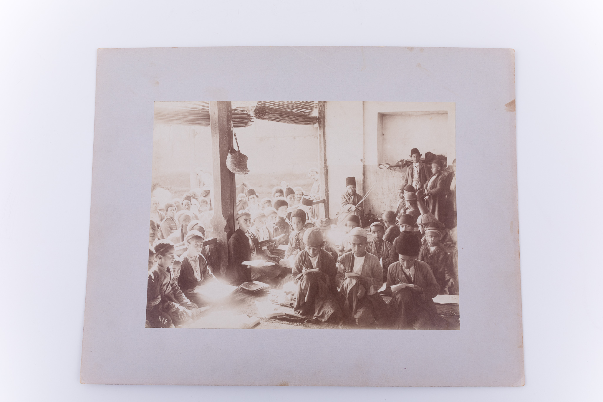 Fotografie de exterior ce prezintă un grup de copii împreună cu învățătorul lor, la școală. Copiii sunt așezați pe jos, cu cărțile în mâini. Pe stradă se remarcă un grup de femei și bărbați. Imaginea este realizată pe hârtie albuminată, după un clișeu pe sticlă și este lipită pe un carton gros, de culoare gri. Pe clișeu, însemnare manuscrisă cu numărul clișeului „879” (practică frecventă în opera lui Antoin Sevruguin). Acceași imagine apare in „Pitt Rivers Museum”, nr. Inv. 2009.7.15. și în „The Nelson Collection of Quajar Photography”. Fotografia din colecția Muzeului ASTRA face parte dintr-un album ce conține 51 de piese, realizate pe hârtie albuminată, aparținând lui Antoin Sevruguin. Acestea prezintă tipuri umane, peisaje și arhitectura din Iran de la sfârșitul secolului al XIX lea și începutul secolului al XX lea, în perioada dinastiei Qajar. Pe coperta albumului, confecționat din imitație de piele roșie, apare textul „Teheran” realizat cu tuș auriu, prin presare, încadrat într-un paspartu cu modele persane, realizat tot prin presare. Pe verso, însemnare manuscrisă, în limba germană, parțial descifrabilă: „Nr. 19. / …”; ștampilă: Muzeul „Brukenthal” Sibiu / Nr. ….; număr de inventar MCPR 2285.