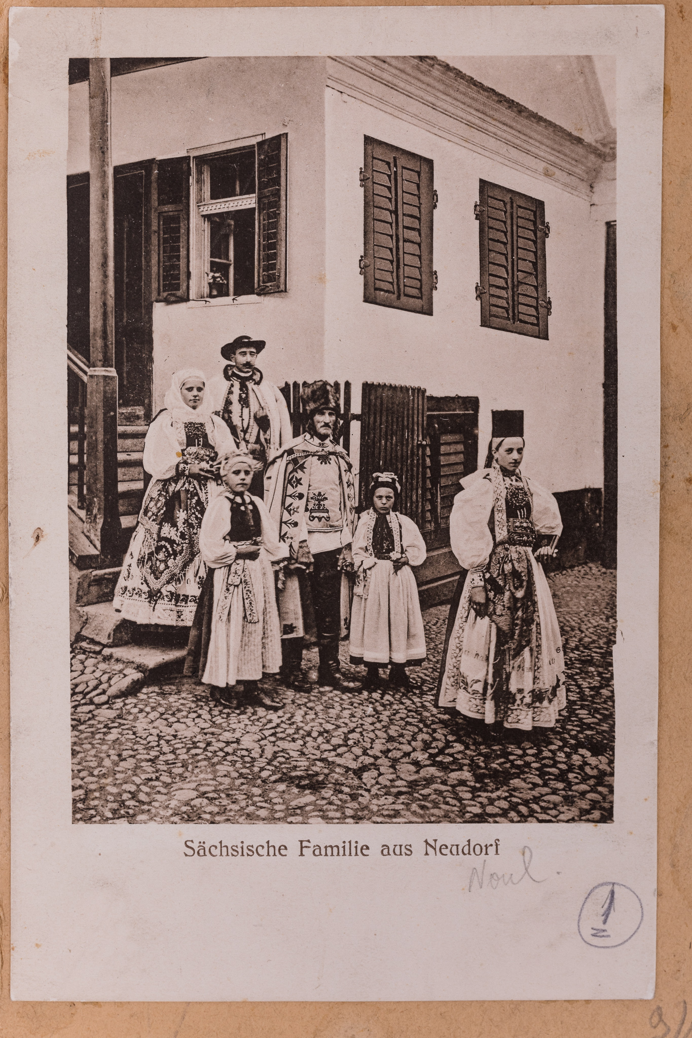 Fotografie, pozitiv după un clișeu pe sticlă, imprimată pe hârtie tipografică, în format carte poștală. Fotografie de exterior ce prezintă o familie în port popular săsesc de sărbătoare din Neudorf, județul Sibiu. Aceștia sunt surprinși în fața locuinței. Sub imagine, text realizat prin imprimare: „Sächsische Familie aus Neudorf.” (Familie de sasi din Noul). Cartea poștală este lipită pe o bucată de carton de culoare crem, alături de alte zece cărți poștale ce prezintă membri ai comunității săsești, în costume de sărbătoare, în diferite ipostaze. Imaginile sunt numerotate, prin însemnare manuscrisă: „1”.