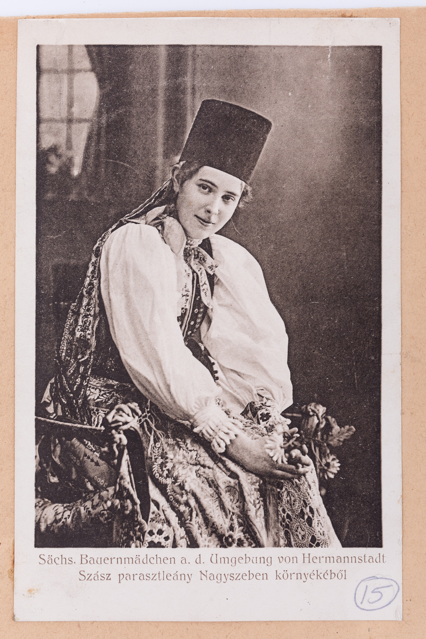 Fotografie, pozitiv după un clișeu pe sticlă, imprimată pe hârtie tipografică, în format carte poștală. Fotografia de atelier cu fundal pictat și elemente de recuzită (soclu de piatră, elemente florale). În prim-plan se află o tânără în costum popular săsesc de sărbătoare din zona Sibiului. Aceasta ține în mâna stângă buchetul de flori. Sub imagine, text realizat prin imprimare: „Säch.Bauernmädchen a.d. Umgebung von Hermannstadt / Szasz paraszleany Nagyszeben kornyekebol” (Tânără săsoaica din jurul Sibiului). Cartea poștală este lipită pe o bucată de carton de culoare crem, alături de alte unsprezece cărți poștale ce prezintă membri ai comunității săsești, în costume de sărbătoare, în diferite ipostaze. Imaginile sunt numerotate, prin însemnare manuscrisă: „15”. În partea de jos a cartonului, ștampilă: „Baron Brukenthal`sches Museum in Hermannstadt” și însemnare manuscrisă: „”Nr. 158./Z. / Berlin.