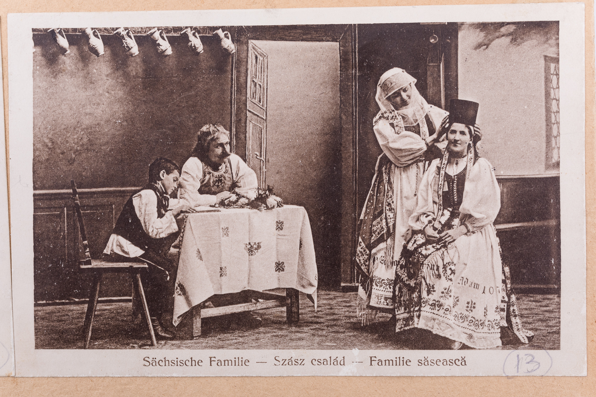 Fotografie, pozitiv după un clișeu pe sticlă, imprimată pe hârtie tipografică, în format carte poștală. Fotografie de atelier, cu fundal pictat și elemente de recuzită (masă, scaune, piese ceramice, piese textile). Prezintă o familie de sași în port popular de sărbătoare. Se remarcă tânăra așezată pe scaun, căreia mama îi aranjează borten-ul (partea a costumului femeiesc săsesc de confirmare). Sub imagine, text realizat prin imprimare în limba germană, maghiară și română: „Sächsische Familie - Szasz csalad - Familie săsească”. Cartea poștală este lipită pe o bucată de carton de culoare crem, alături de alte douăsprezece cărți poștale ce prezintă membri ai comunității săsești, în costume de sărbătoare, în diferite ipostaze. Imaginile sunt numerotate, prin însemnare manuscrisă: „13”. În colțul din stânga jos al cartonului, ștampilă: „Baron Brukenthal`sches Museum in Hermannstadt”.