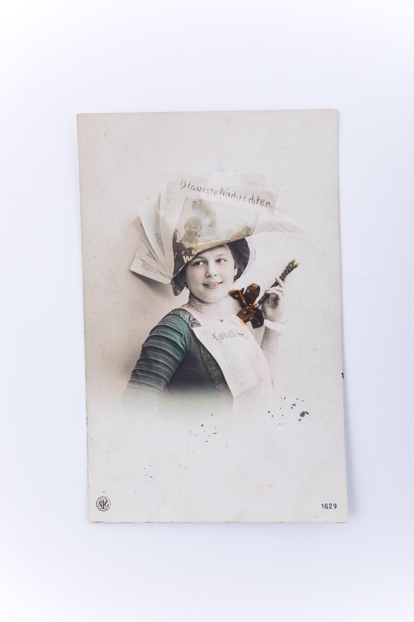 Cromolitografie imprimată pe hârtie tipografică în format carte poștală. Prezintă o tânără, într-o fotografie tip medalion, în costum orășenesc de început de secol XX. Tânăra are pe cap o pălărie realizată din hârtie de ziar cu text în limba germană. O altă bucată de ziar este prinsă pe piept. Pe pălărie se distinge un titlu de ziar „Blaueste Nachrichten” (traducere, potrivit limbii germane vechi, „Veștile uimitoare”), iar pe piept, se distinge numele ziarului: „Lokal”. În partea de jos a imaginii, prin imprimare marca editorului, încadrată într-un cerc: „N.P.G.” (Neue Photographische Gesellschaft), iar în colțul din stânga apare codul de identificare al cărții poștale: „1629”. Modelele aurii de pe rochia tinerei sunt evidențiate prin colorare manuală. Verso: rubricatură de carte poștală; text greu descifrabil, în limba germană. Se distinge data la care a fost trimisă cartea poștală, atât pe ștampila poștală aplicată peste timbru, cât și în textul propriu-zis: 30 iunie 1911. Text în limba germană, greu descifrabil și semnătura expeditorului: „Kessler”. Destinatarul acestei cărți poștale este „Frauleine Tilli Zilg”. Adresa de expediere: „Loco / Langgasse”. Etichetă albă cu numărul de inventar: MCPT 2682.