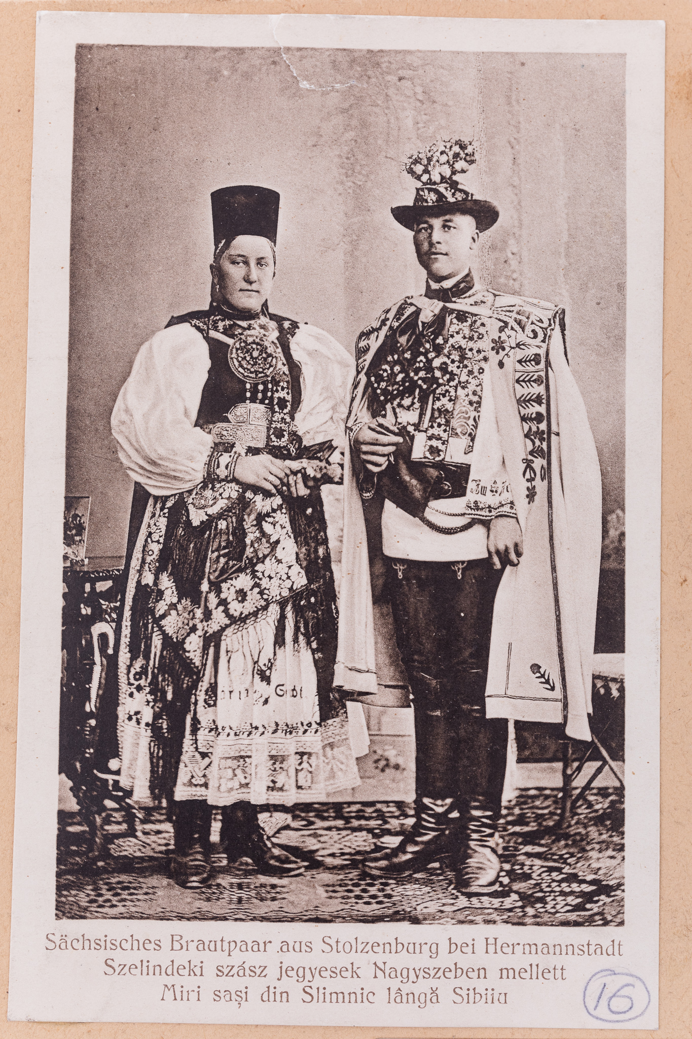 Fotografie, pozitiv după un clișeu pe sticlă, imprimată pe hârtie tipografică, în format carte poștală. Fotografia de atelier cu fundal pictat și elemente de recuzită (scaun, masă). În prim-plan se află o tânără pereche în costume populare săsești de sărbătoare din Slimnic, județul Sibiu. Sub imagine, text realizat prin imprimare: „Sächsisches Brautpaar aus Stolzenburg bei Hermannstadt / Szelindeke szasz jegyesek Nagyszeben mellett / Miri sași din Slimnic lângă Sibiu”. Cartea poștală este lipită pe o bucată de carton de culoare crem, alături de alte unsprezece cărți poștale ce prezintă membri ai comunității săsești, în costume de sărbătoare, în diferite ipostaze. Imaginile sunt numerotate, prin însemnare manuscrisă: „16”. În partea de jos a cartonului, ștampilă: „Baron Brukenthal`sches Museum in Hermannstadt” și însemnare manuscrisă: „”Nr. 158./Z. / Berlin.