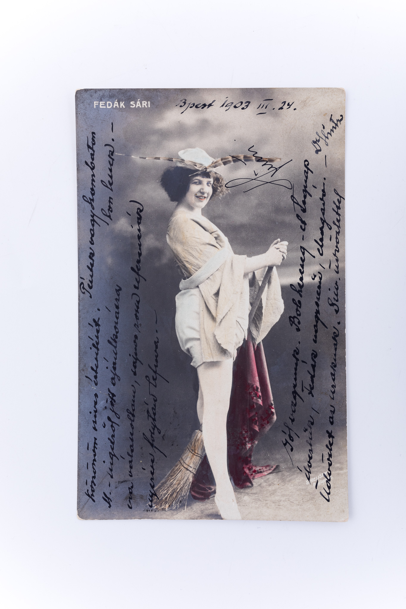 Cromolitografie imprimată pe hârtie tipografică în format carte poștală. O prezintă pe actrița Fedak Sari, în costum de scenă, într-o fotografie de atelier cu fundal pictat și elemente de recuzită: mătură. Text și semnătură indescifrabile, în limba maghiară, localizare și datare: Budapesta, 1903, 24 martie. Imaginea a fost colorată manual: mantia, mătura. Verso: rubricatură de carte poștală; text greu descifrabil, în limba maghiară. Text imprimat, în 4 limbi (germană, franceză, maghiară, engleză): „Postkarte - Correspondenzkarte” și ștampilă cu data și localitatea expedierii: „1903. -MAR 24. / Budapest”. Localitatea în care a fost trimisă cartea poștală este Cluj-Napoca. În colțul din stânga sus apare ștampila oficiului poștal din Cluj-Napoca și data: 25 martie 1903. Etichetă albă cu numărul de inventar: MCPT 2682.