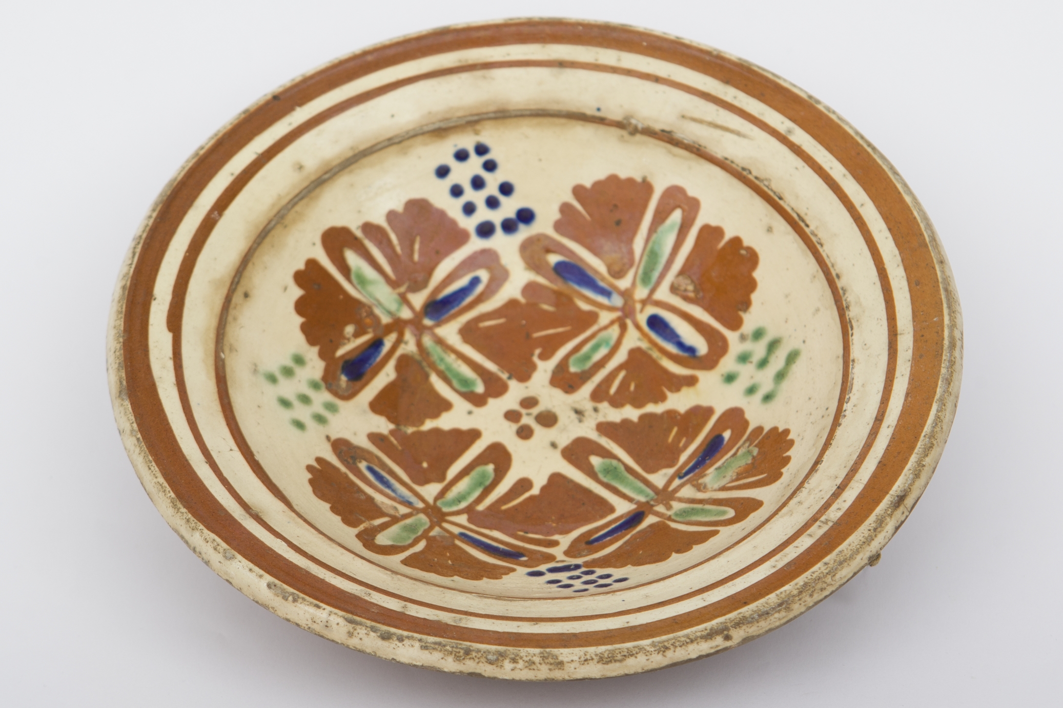 Piesă ceramică smălțuită la interior, de formă tronconică, cu baza plată, pereții înalți și marginea evazată. Peste stratul crem de angobă a fost pictat decorul geometrizant și fitomorf stilizat. Pe fundul vasului au fost realizate patru flori maro, cu mijlocul verde și albastru, fiind mărginite de patru grupuri de puncte verzi și albastre (două câte două, în diagonală). Marginal, pe buza piesei au fost realizate trei linii drepte circulare, ultima fiind mai groasă spre deosebire de primele două. Farfuria era folosită în gospodărie pentru decorarea interioarelor. Prezintă uzură funcțională. Cromatică: fond: cărămiziu; crem; motive: maro; verde; albastru.
